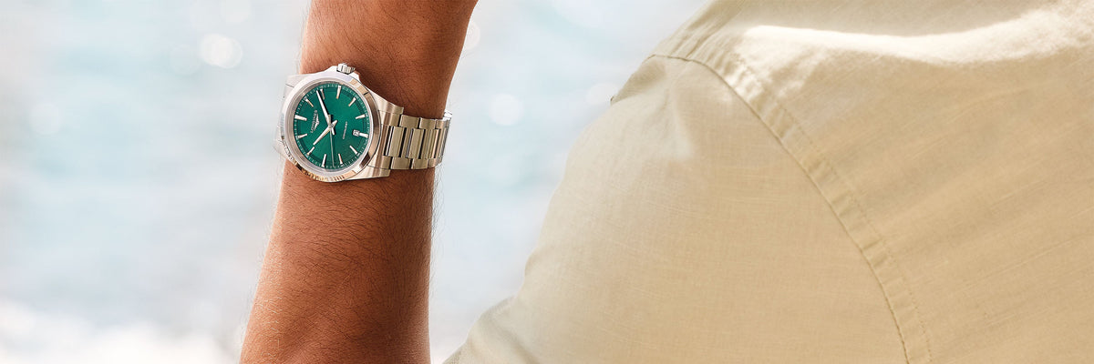 Eine Longines-Uhr mit grünem Zifferblatt und silbernem Stahlarmband am Handgelenk eines Mannes, der ein beiges Hemd trägt, vor einem verschwommenen Meereshintergrund.