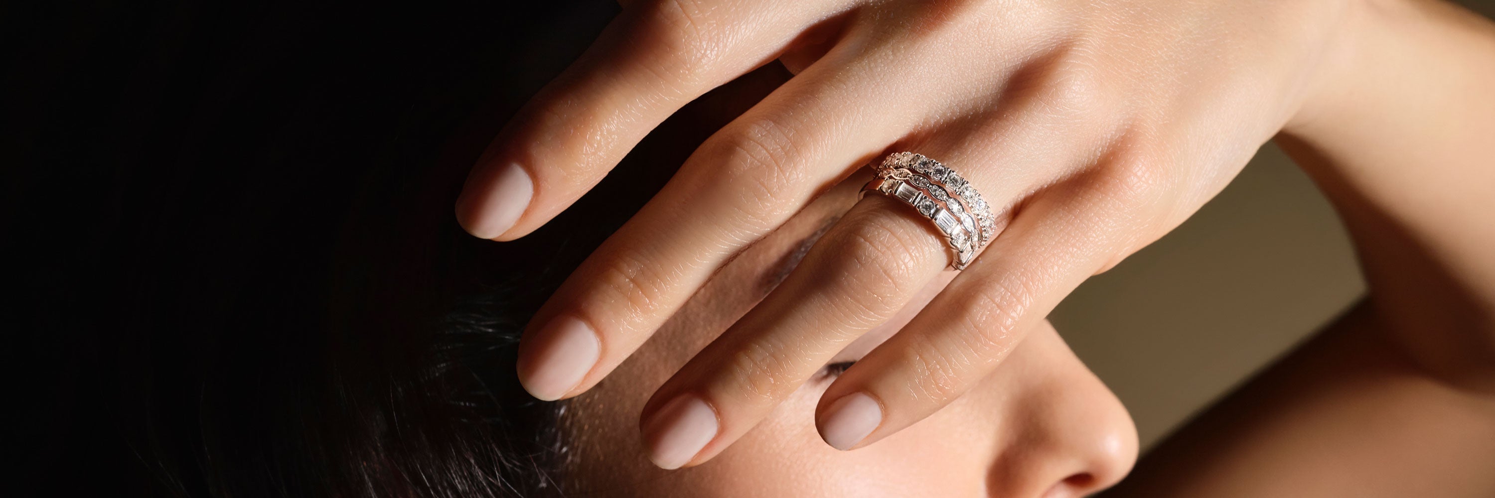 Frau in lockigem Haar blickt hinter einer Wand hervor, auf dieser ihre Hand liegt. An ihrem Mittelfinger trägt sie einen mit Steinen besetzten Ring der Kollektion Memoire des Schmuckherstellers Schullin.