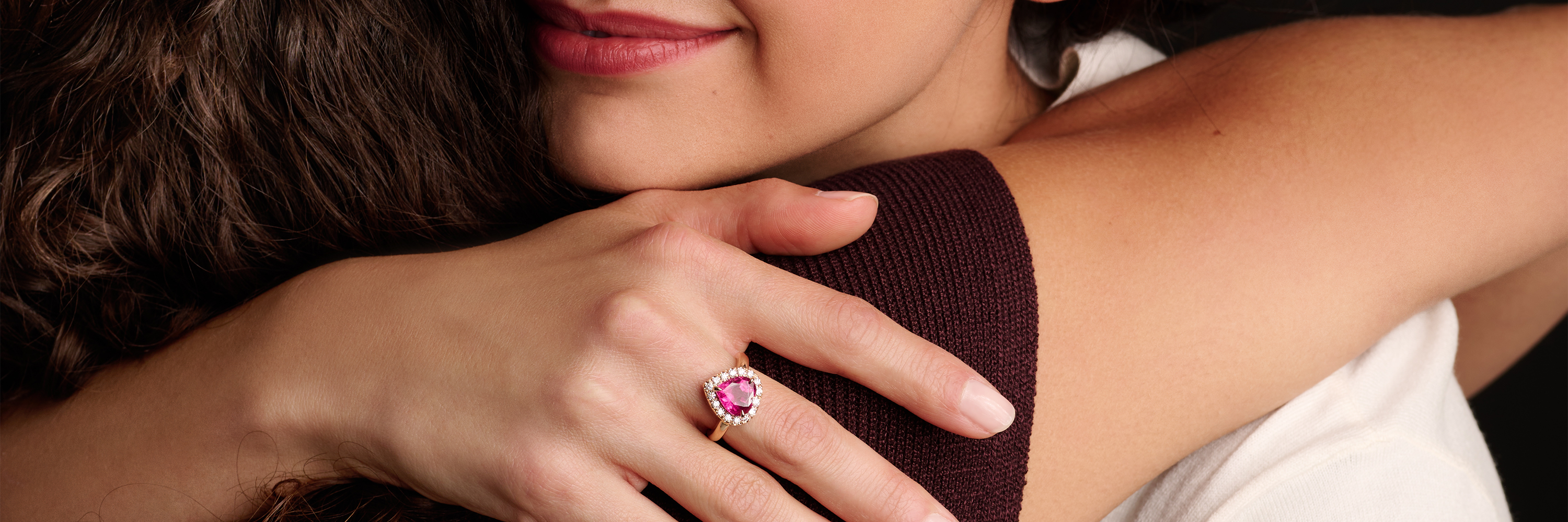 Eine Frau mit dunklem Haar und rosafarbenen Edelsteinohrringen legt ihr Gesicht sanft an den Kopf einer blonden Frau, beide mit nackten Schultern vor einem neutralen Hintergrund.