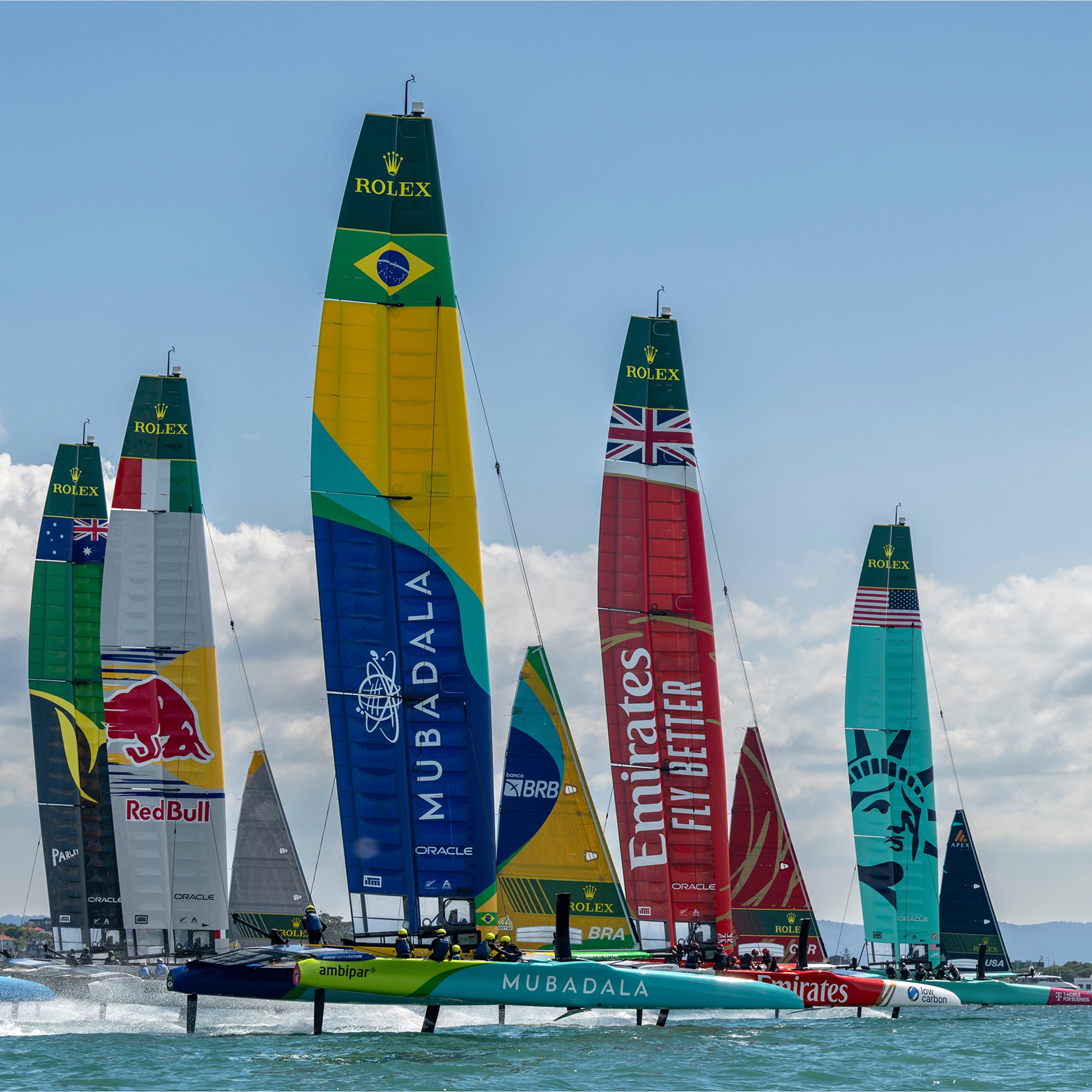 Farbenfrohe Segelboote mit Markensegeln, darunter Red Bull, Mubadala, Emirates und eine Freiheitsstatue, fahren auf dem Wasser unter einem blauen Himmel mit vereinzelten Wolken.