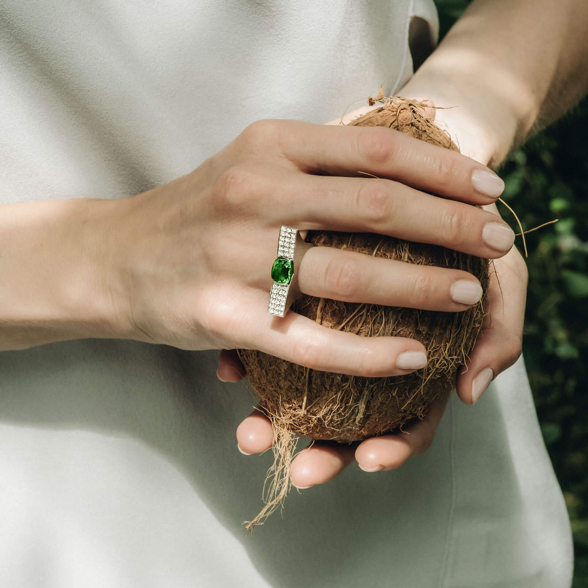 Eine Person hält eine Kokosnuss in beiden Händen und präsentiert den TSAVORIT RING GHIBELLINI von Schullin an ihrem rechten Zeigefinger. Dieser exquisite Ring zeigt einen großen Tsavorit-Edelstein, der von kleineren Diamanten umgeben ist, alles in 18 Karat Weißgold gefasst. Der verschwommene Hintergrund aus natürlichem Grün verstärkt die bezaubernde Szene.