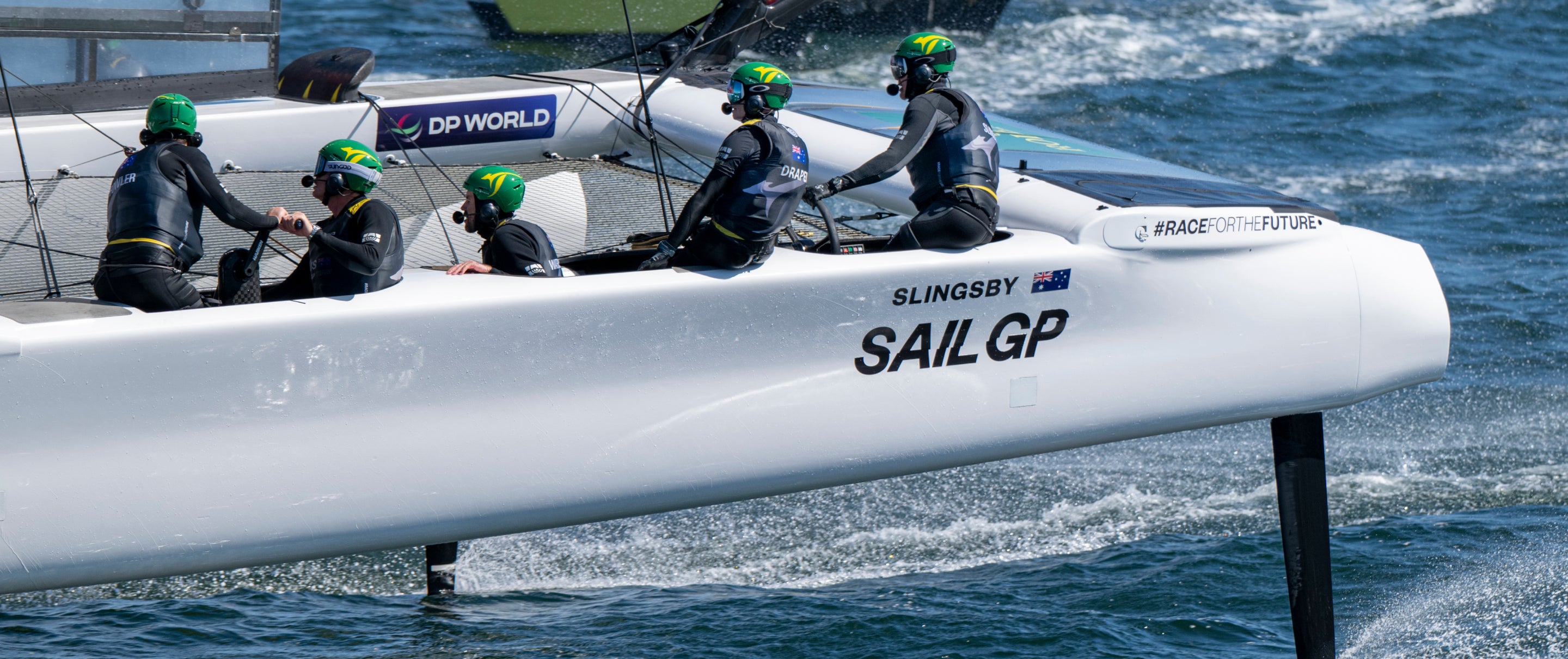 Vier Segler mit Helmen und Neoprenanzügen fahren auf einem weißen Hochgeschwindigkeits-Katamaran mit der Aufschrift "SLINGSBY SAIL GP" um die Wette, der mit sichtbaren Tragflügeln über das Wasser gleitet.