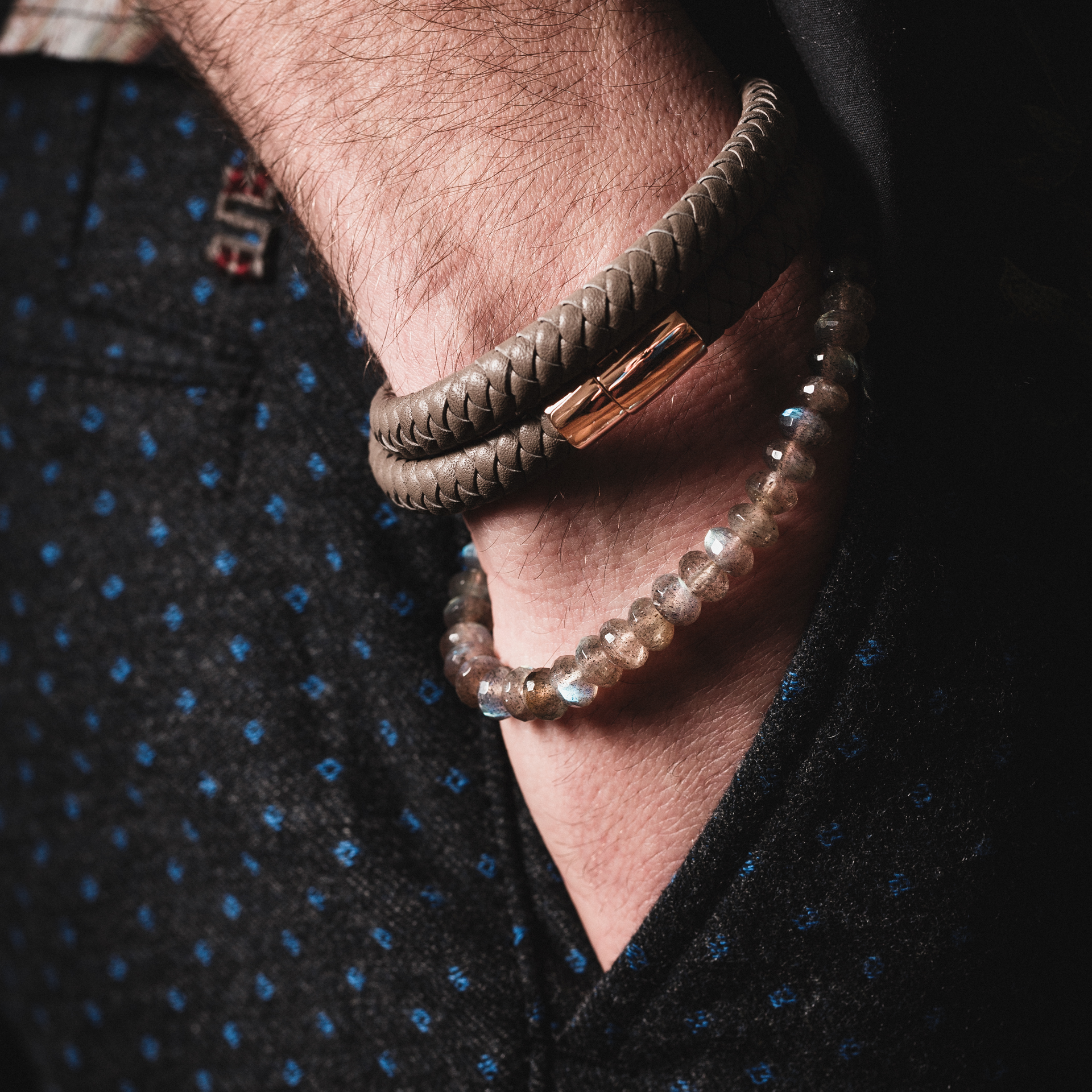 Nahaufnahme der Hand einer Person in einer Tasche, die zwei Armbänder zeigt: das POWER ARMBAND von Schullin aus geflochtenem Taupe-Leder mit einem Verschluss aus 18-karätigem Roségold und ein Perlenarmband mit schillernden Steinen. Die Person trägt dunkle, gemusterte Hosen.