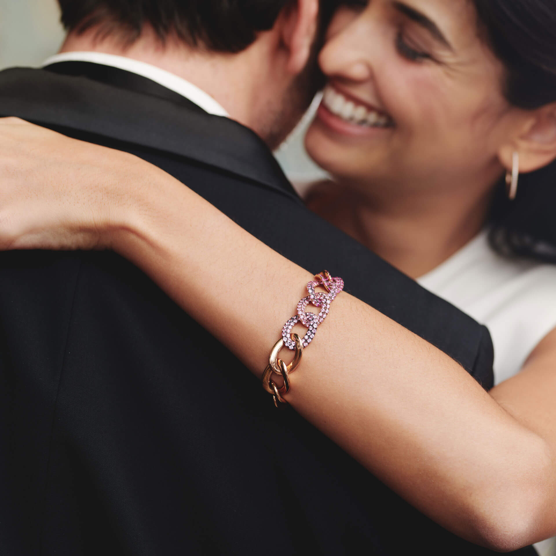 Eine Frau im weißen Kleid umarmt einen lächelnden Mann im Anzug. Sie trägt das atemberaubende SAPHIR ARMBAND PINK von Schullin mit ineinandergreifenden Gliedern. Der Fokus liegt auf ihrem Handgelenk und dem Armband, das den zärtlichen Moment einfängt.