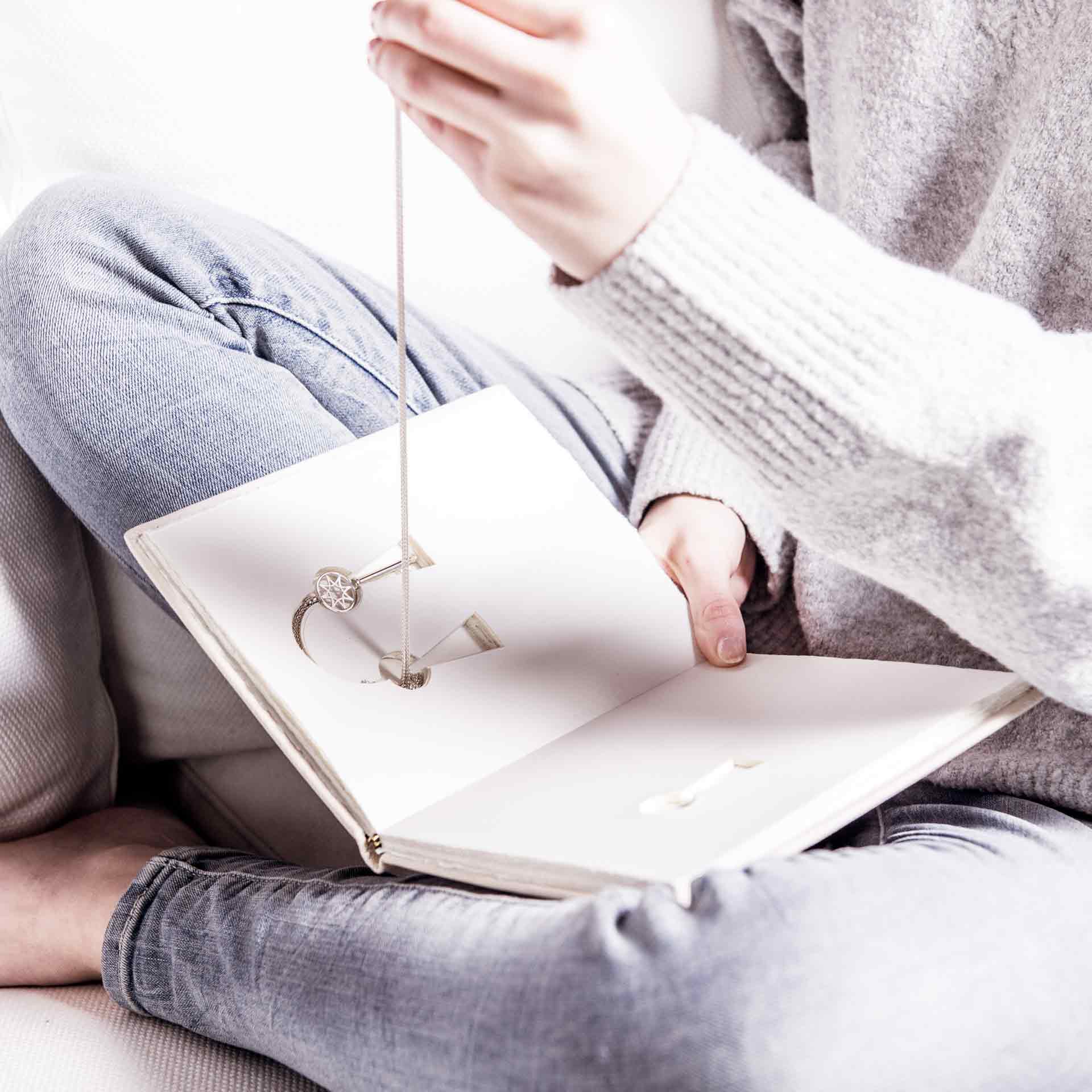 Eine Person sitzt im Schneidersitz und hält ein offenes Buch mit dem SCHLÜSSEL ZUM GLÜCK KETTE & BUCH von Schullin darin. Das Buch hat ein Fach, in das die dreireihige Kette mit einem runden Anhänger passt. Die Person trägt hellgraue Kleidung und Jeans.