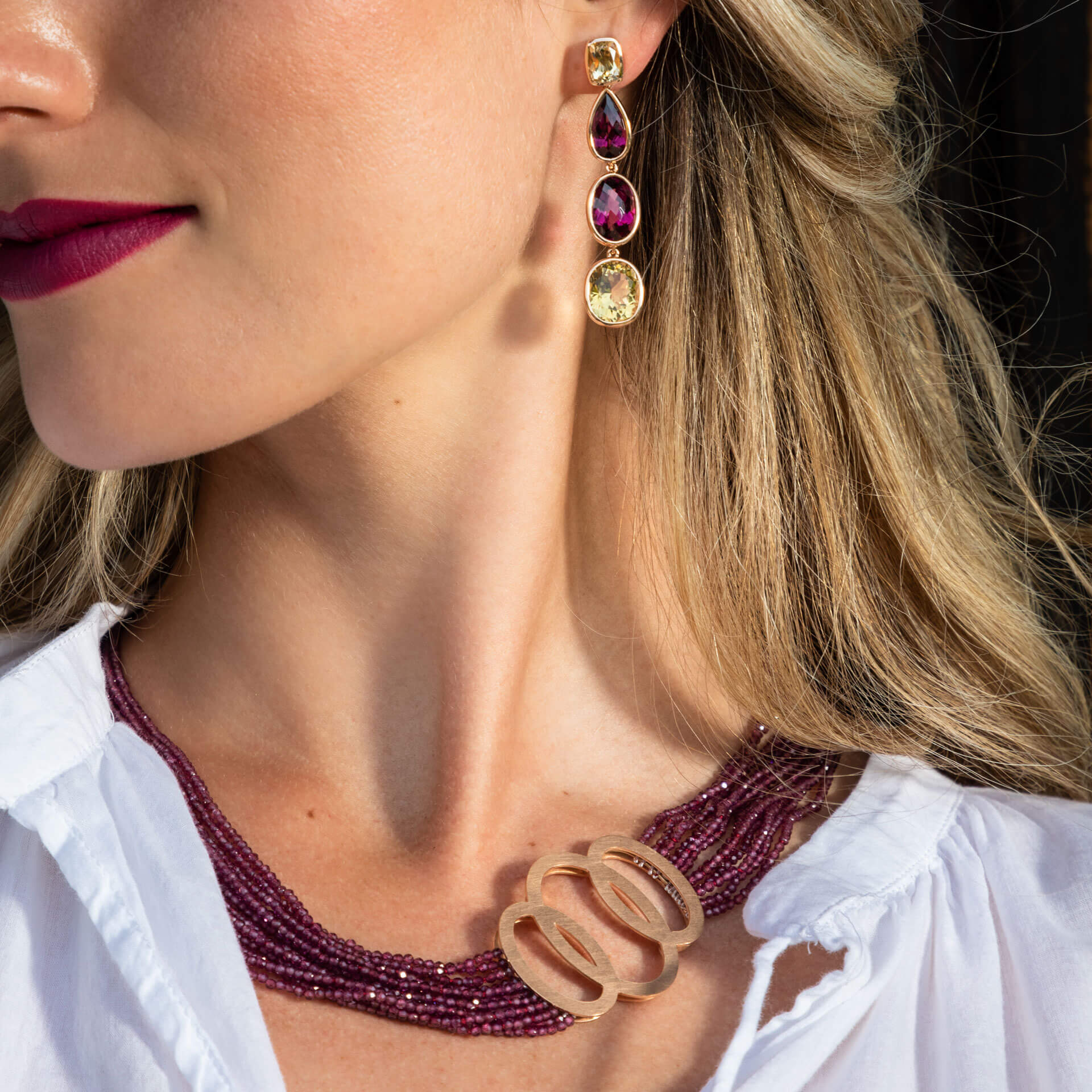 Eine Person, geschmückt mit einer mehrreihigen, dunkelroten Perlenkette mit dekorativem Rundverschluss, gepaart mit den eleganten RHODOLIT OHRGEHÄNGE von Schullin. Das Ensemble wird durch leuchtend gelbe, rosa und tiefrote Edelsteine in den Ohrringen mit Rhodolith-Highlights auf einer strahlend weißen Oberseite akzentuiert.