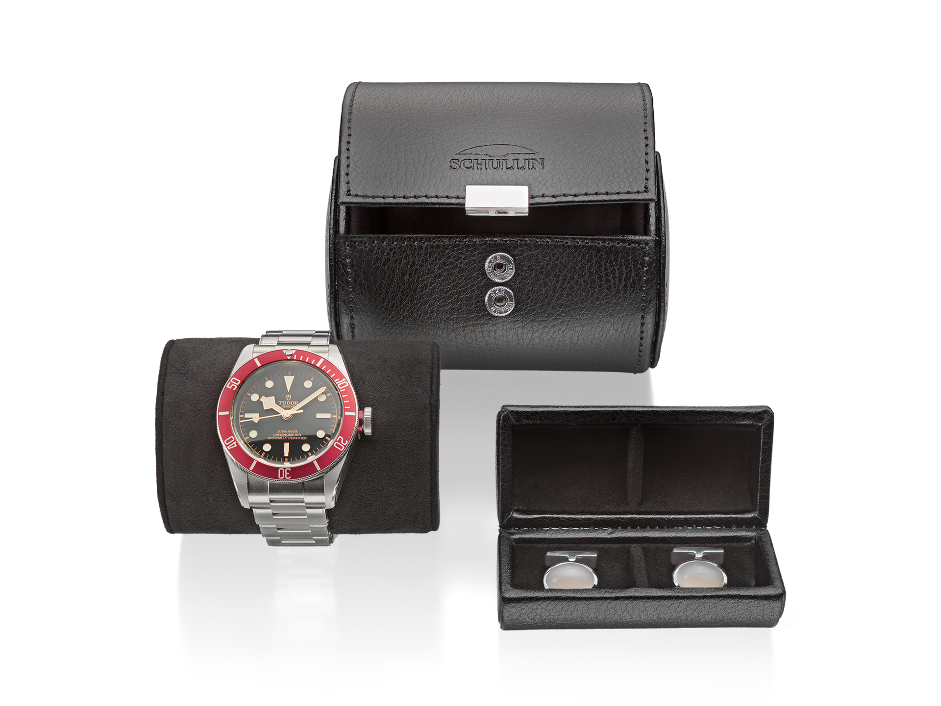 Eine silberne Armbanduhr mit roter Lünette und schwarzem Zifferblatt wird elegant auf einem schwarzen Kissen präsentiert. Daneben befindet sich eine elegante UHRENROLLE von Schullin, die das Display perfekt in Szene setzt. Daneben enthält eine kleinere Box exquisite silberne Manschettenknöpfe.
