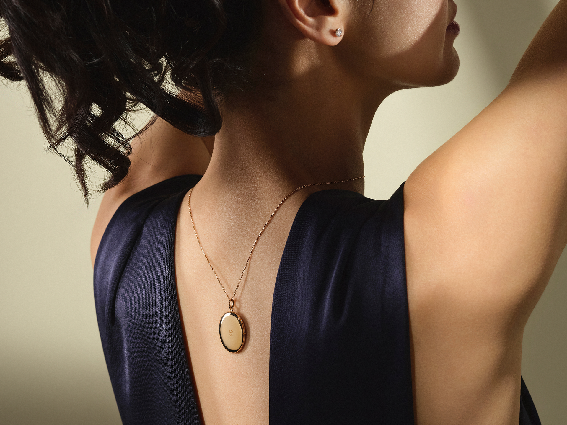 Eine Frau mit hochgestecktem Haar trägt die goldene Halskette MEDAILLON LOVE von Schullin und einen kleinen Ohrstecker, gepaart mit einem ärmellosen dunklen Kleid, von hinten vor einem neutralen Hintergrund gezeigt.