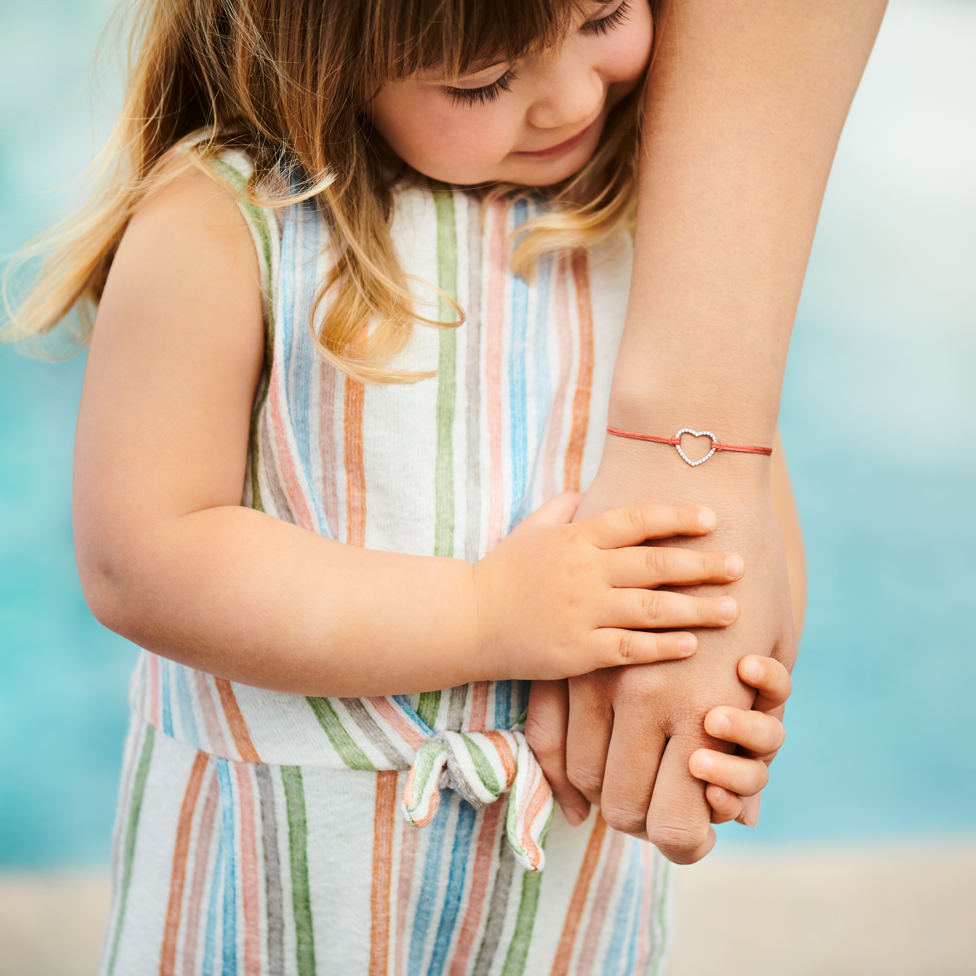 Ein kleines Kind in einem gestreiften Kleid umarmt den Arm eines Erwachsenen, der mit dem Schullin HERZ ARMBAND WEISSGOLD mit einem roten Herzanhänger geschmückt ist, während das Kind vor einem blauen, verschwommenen Hintergrund süß lächelt.