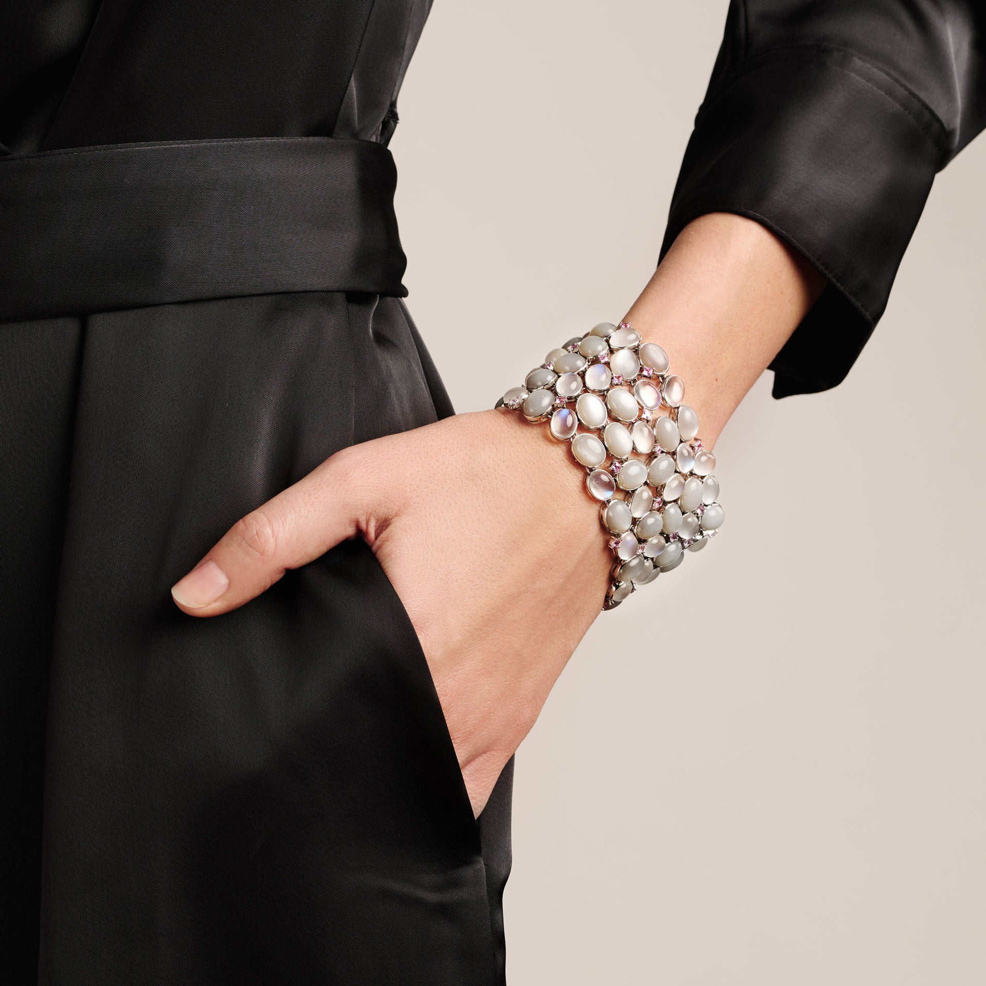 Eine Person in einem schwarzen Satinkleid hat eine Hand in der Tasche und hebt das Schullin MONDSTEIN ARMBAND hervor, ein breites und elegantes Armband, das mit mehreren ovalen Silbersteinen besetzt ist.