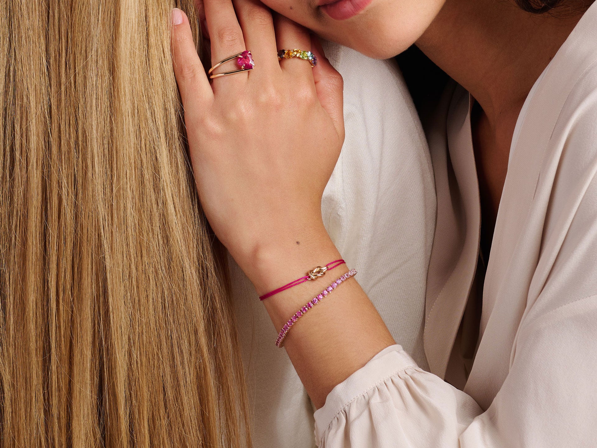 Eine Frau mit langen blonden Haaren stützt ihren Kopf auf die Schulter eines anderen, sie trägt eine weiße Bluse und das SAPHIR TENNISARMBAND von Schullin sowie goldene Ringe mit rosa Steinen. Das Bild hebt ihre Hand und ihren Schmuck hervor.