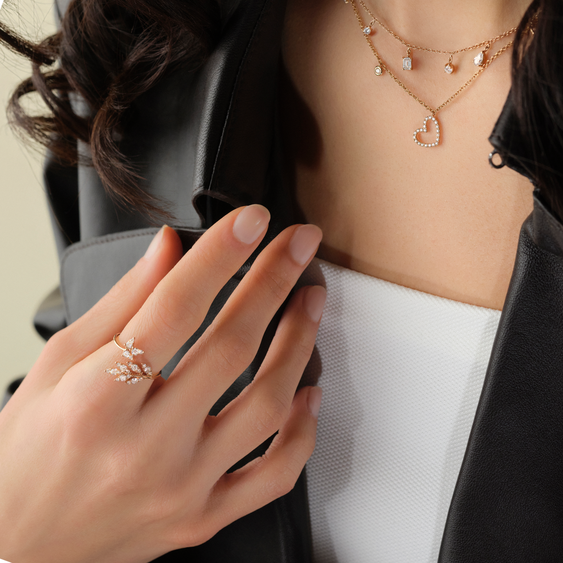 Eine Frau in schwarzer Lederjacke und weißem Oberteil modelliert die HERZ HALSKETTE von Schullin, eine goldene Lagenkette mit einem Herzanhänger, zusammen mit einem Ring in Blattgoldoptik.
