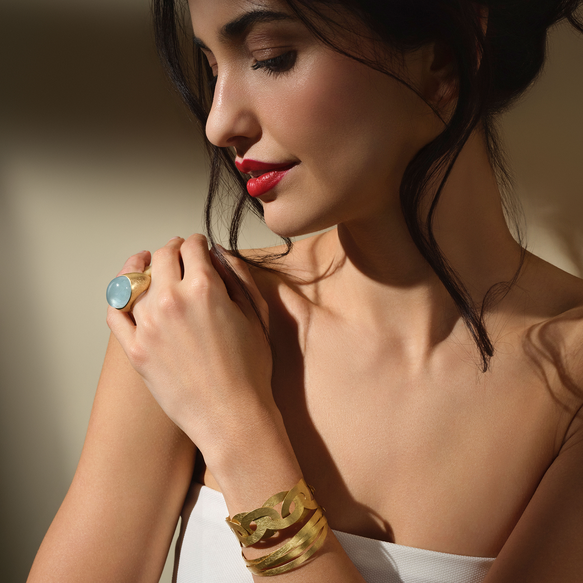 Eine Frau mit dunklem Haar und rotem Lippenstift blickt nach unten. Sie trägt ein weißes trägerloses Oberteil, ein breites Armband aus 18 Karat Gelbgold und den Schullin AQUAMARIN RING BUBBLE, wobei ihr Gesicht und ihr Schmuck in sanftem Licht erstrahlen.