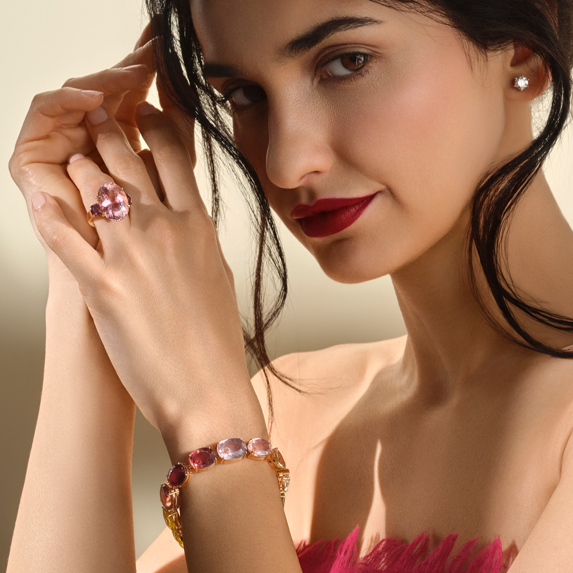 Eine Frau mit dunklem Haar und rotem Lippenstift trägt den Schullin TURMALIN RING, ein rosafarbenes, herzförmiges Stück, zusammen mit einem mehrfarbigen Edelsteinarmband und einem sichtbaren Diamantohrstecker.