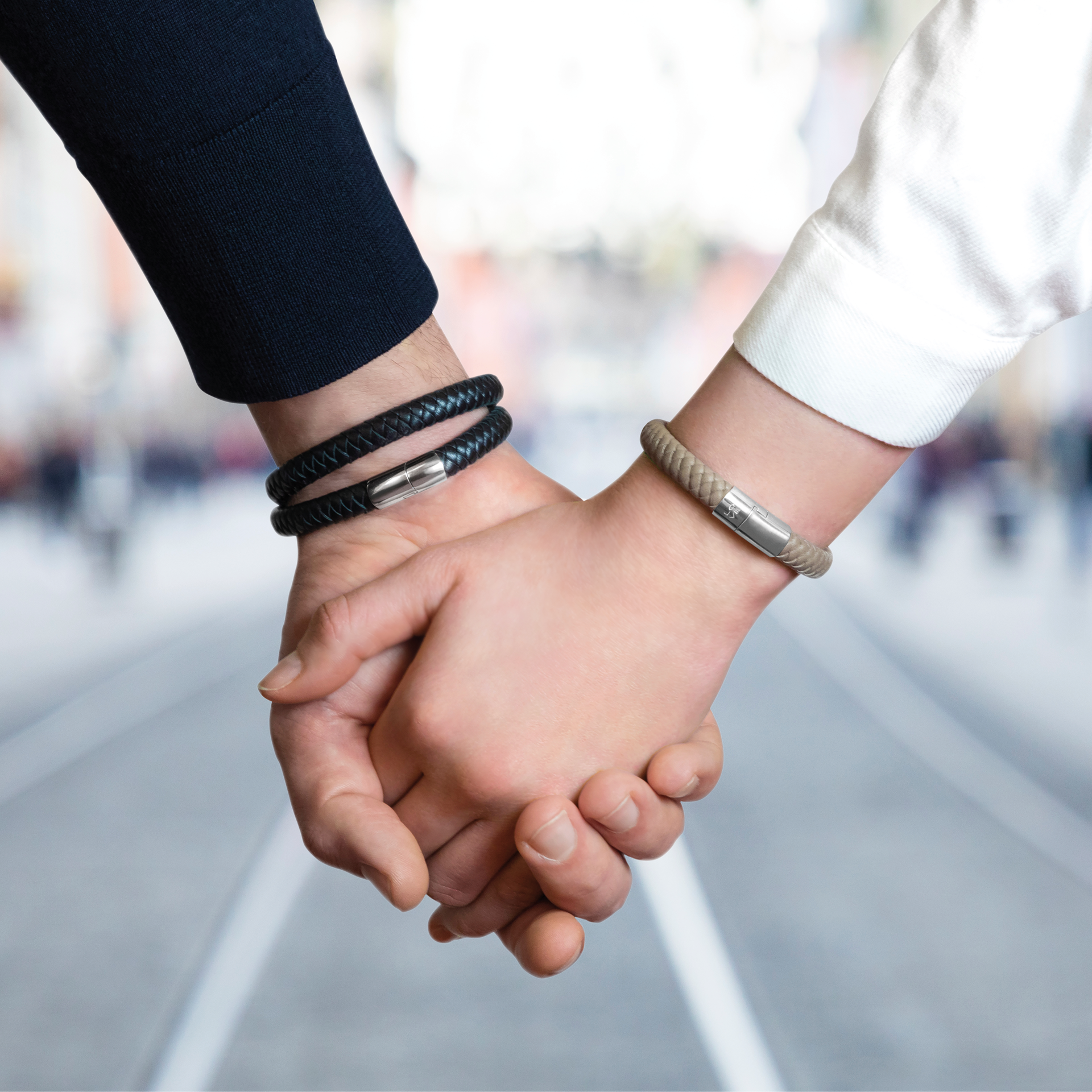 Zwei Personen halten sich an den Händen und tragen Schullin POWER ARMBAND STERLING SILBER - eine mit einer silbernen Schließe, die andere mit einem Lederband - vor einer verschwommenen Stadtstraße und Straßenbahnschienen im Hintergrund.
