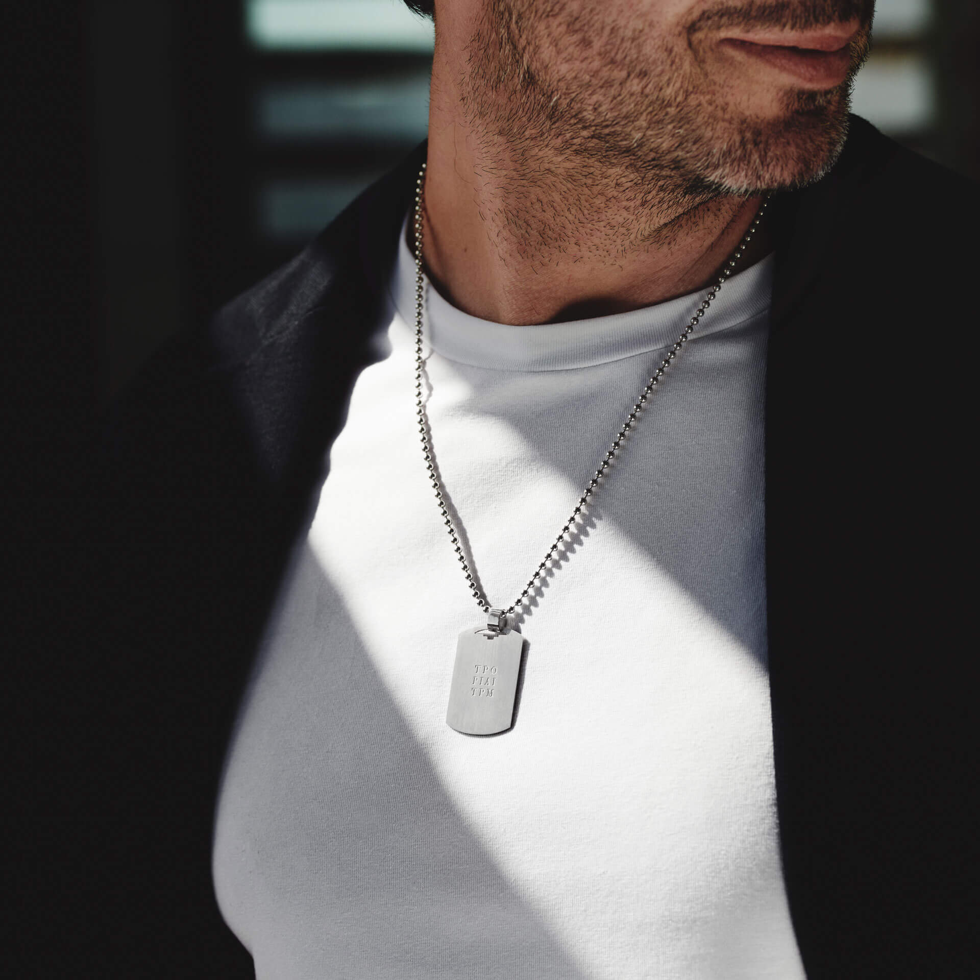 Ein Mann ist von den Schultern abwärts abgebildet. Er trägt ein weißes T-Shirt und eine dunkle Jacke und einen kurzen Bart. Als Accessoire trägt er die Schullin ID HALSKETTE aus Sterlingsilber, die eine gravierte Plakette mit der Gravur „Let God“ aufweist, die in der sonnenbeschienenen Umgebung schimmert.