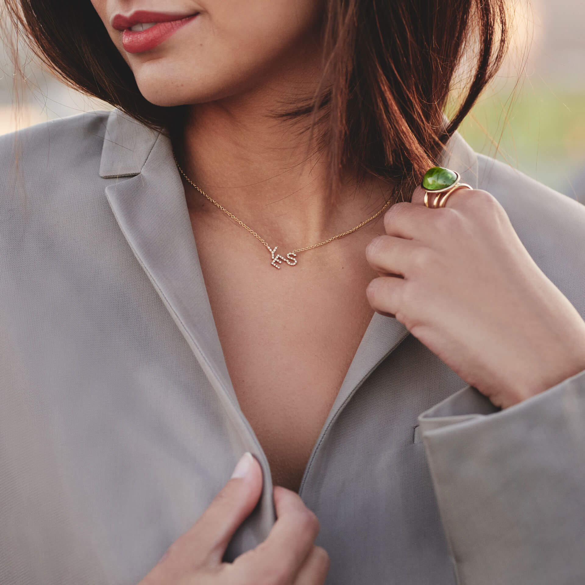 Eine Frau in einem beigen Blazer trägt die YES HALSKETTE von Schullin, eine Halskette aus 18 Karat Gelbgold mit Initialen. Sie kombiniert sie mit einem großen Ring mit grünem Edelstein und hält sanft das Blazerrevers, wodurch ihre atemberaubenden Accessoires vor einem verschwommenen Hintergrund hervorgehoben werden.