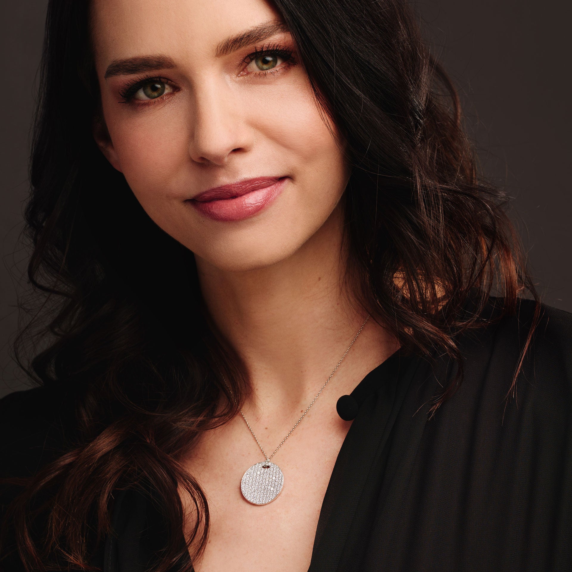 Eine Frau mit langen dunklen Haaren und hellen Augen, in einem schwarzen Oberteil, lächelt sanft, während sie die Schullin DIAMANT HALSKETTE in 18 Karat Weißgold trägt, die in einem Studio-Porträt vor einem dunklen Hintergrund zu sehen ist.