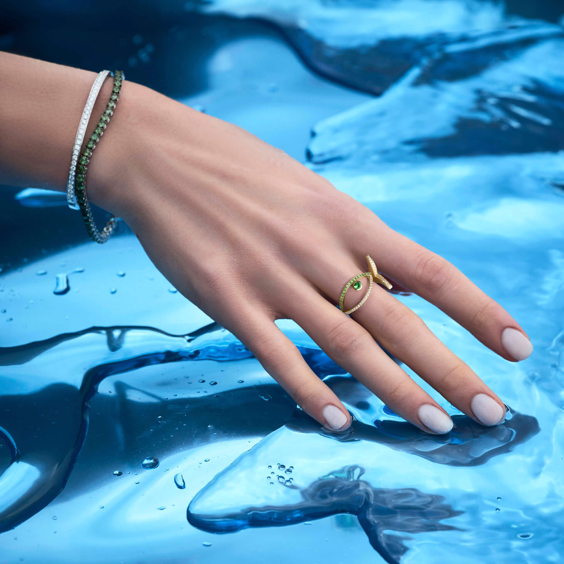 Eine Hand mit manikürten Nägeln ruht auf einer glänzenden blauen Oberfläche, geschmückt mit einem 18 Karat Gelbgoldarmband und dem Schullin TSAVORIT RING FISH. Der Hintergrund ähnelt schmelzendem Eis und schafft eine glatte, reflektierende Umgebung.