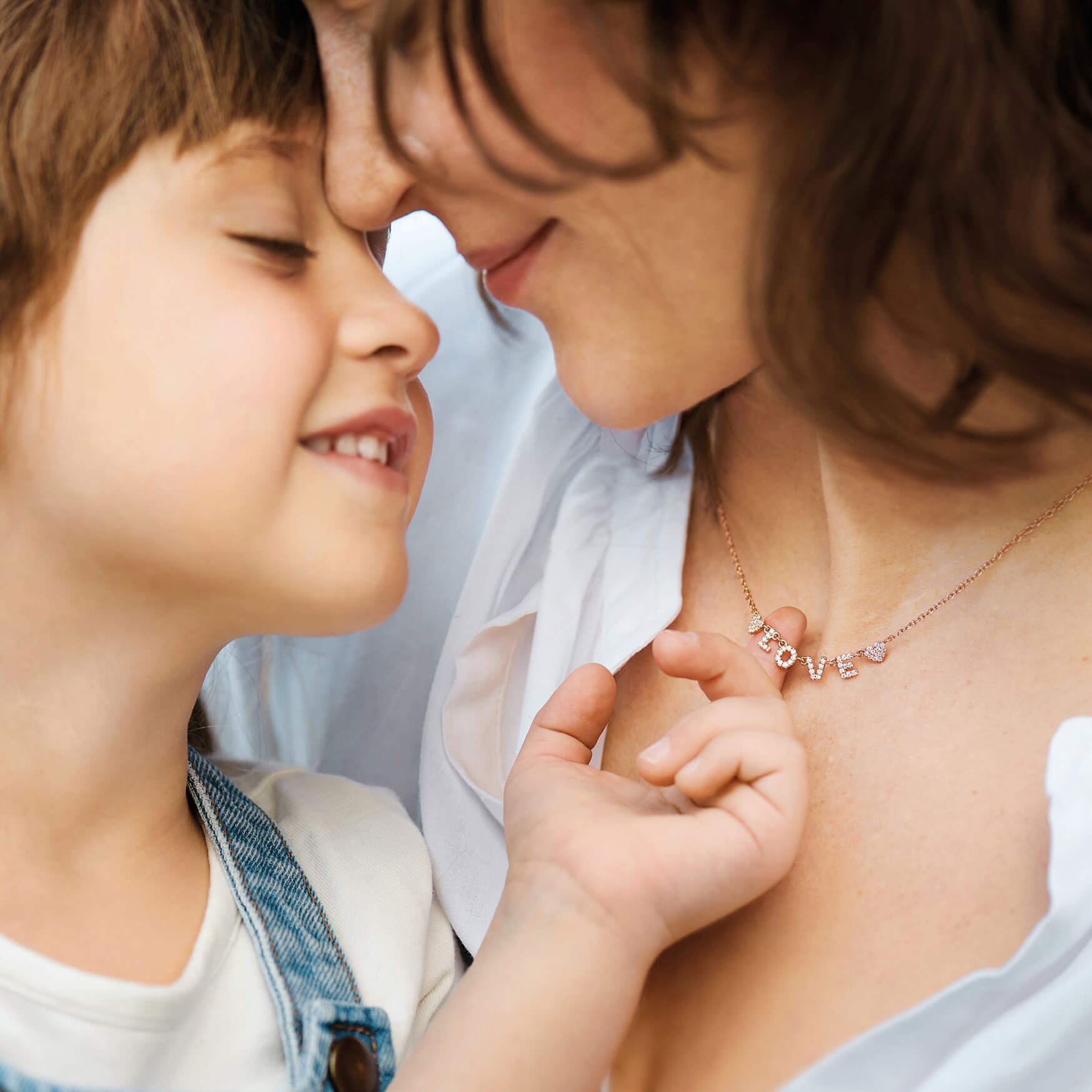 Eine Nahaufnahme einer Frau und eines Kindes, die einen zärtlichen Moment teilen, wobei sich ihre Nasen berühren und ihre Augen geschlossen sind. Die Frau trägt Schullins roségoldfarbene LOVE HALSKETTE. Das Kind trägt ein weißes Hemd und einen Jeans-Overall und fängt damit die perfekte Essenz eines Muttertagsgeschenks ein.