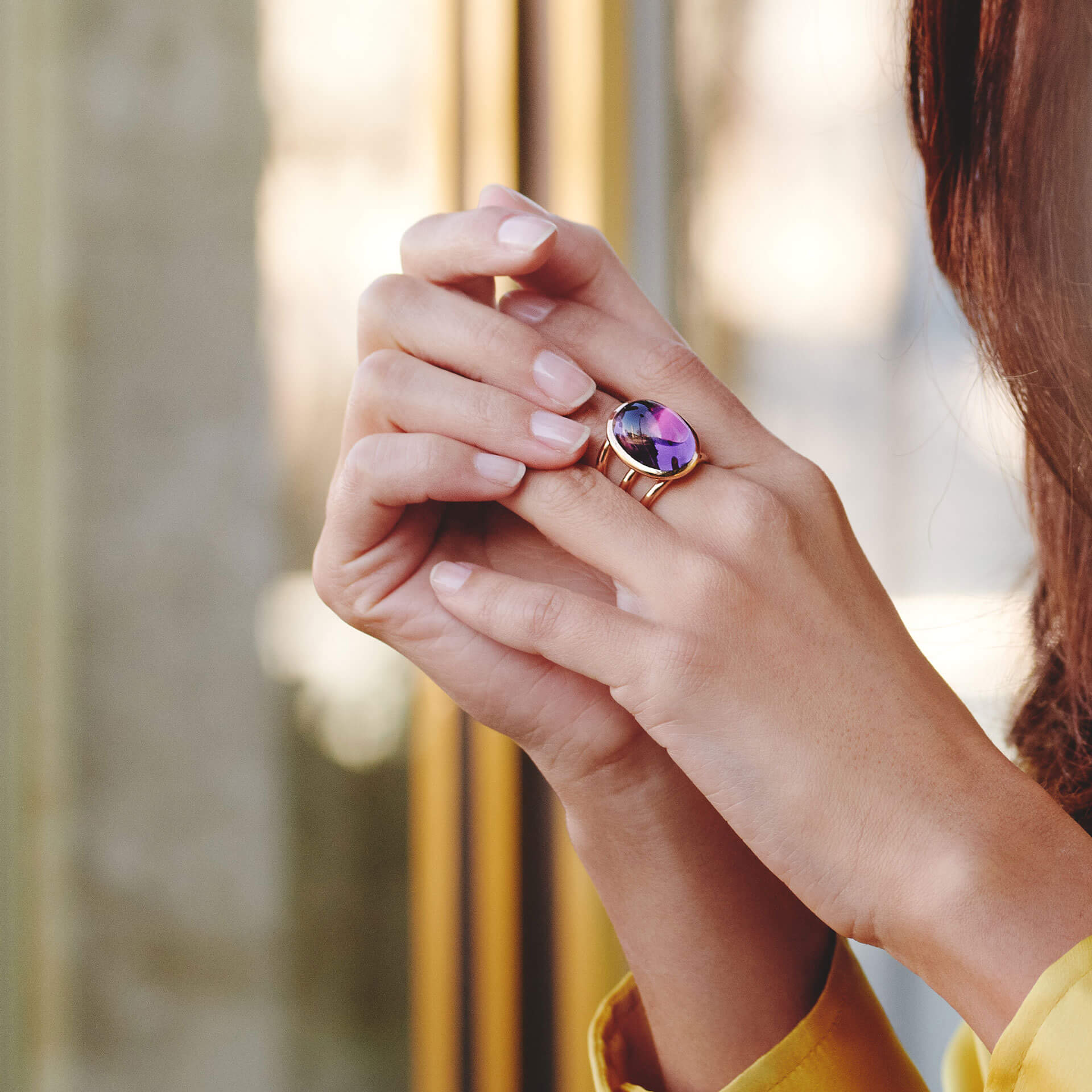 Eine Person, die den Schullin AMETHYST RING TRINITY mit einem großen, runden Amethyst-Edelstein in Roségold trägt, hält ihre Hand sanft vor einer reflektierenden Glasoberfläche. Heller Nagellack ergänzt die Szene und der sanft verschwommene Hintergrund unterstreicht die Eleganz dieses exquisiten Schmuckstücks.