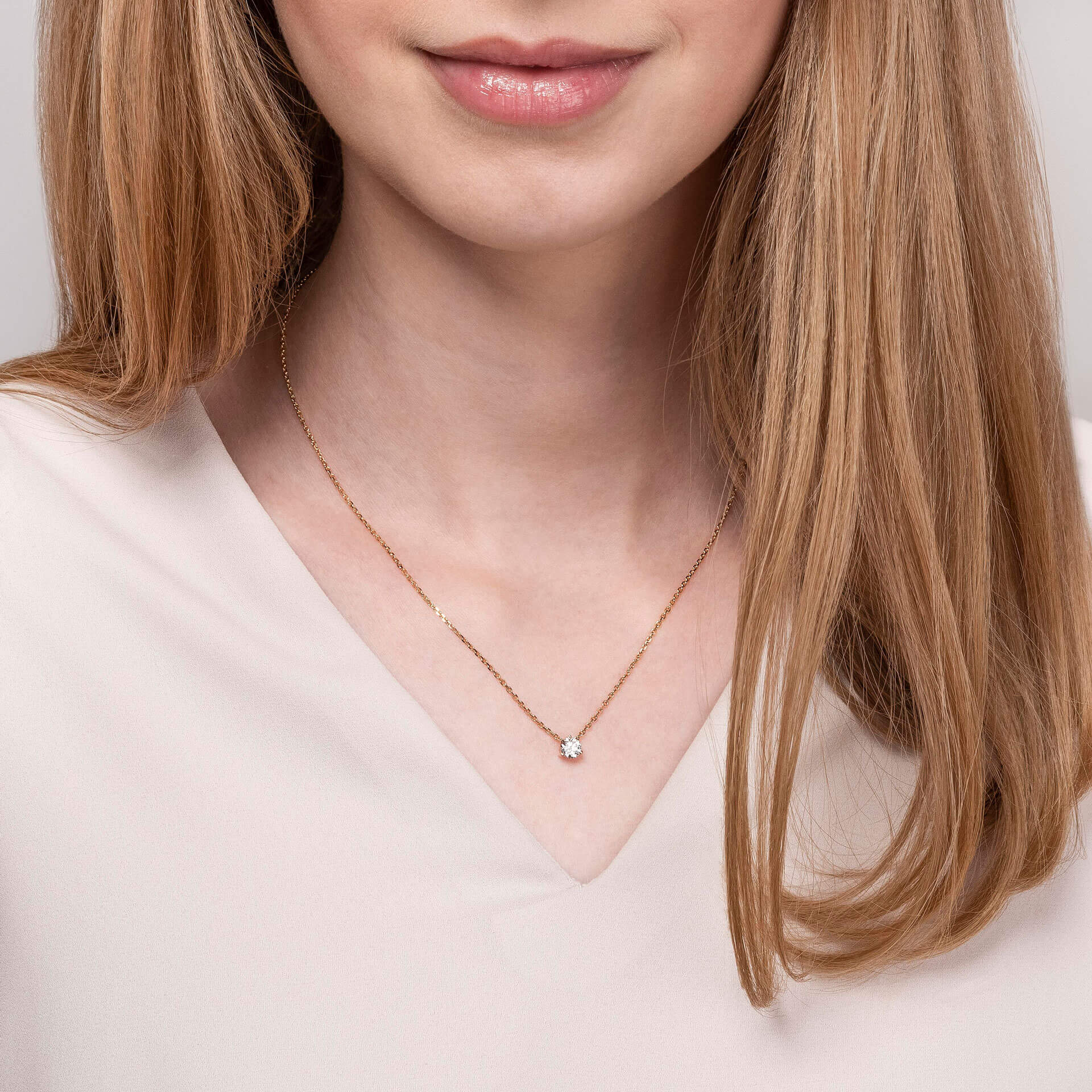 Eine Frau mit hellbraunem Haar trägt ein blassrosa Oberteil mit V-Ausschnitt und die Schullin SOLITAIRE HALSKETTE 0,45 CARAT in Gold mit einem kleinen runden Anhänger. Das Bild ist von den Schultern bis knapp über die Lippen abgeschnitten.