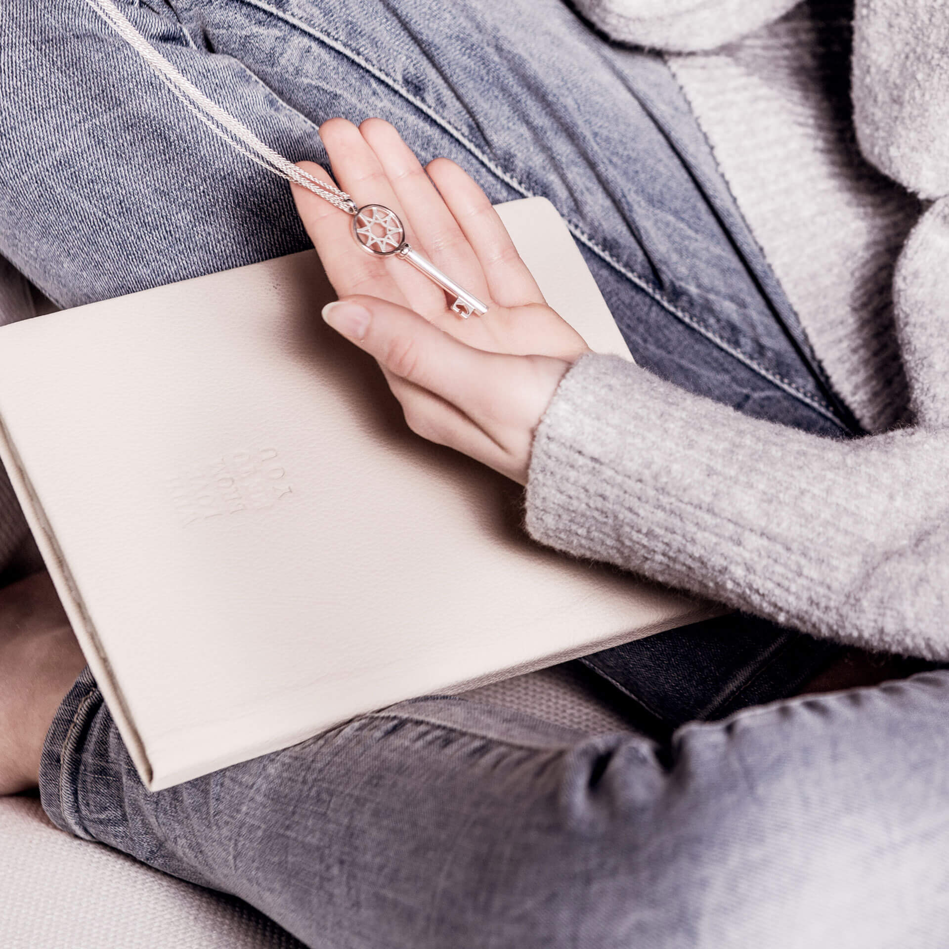 Eine Person in grauem Pullover und Jeans hält die Halskette SCHLÜSSEL ZUM GLÜCK von Schullin, deren Sterling-Silber-Anhänger sanft schimmert, während das dazugehörige beige Hardcover-Buch auf ihrem Schoß liegt.