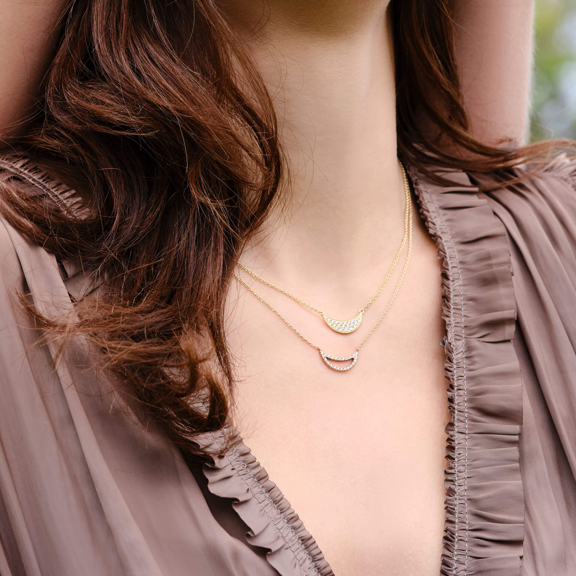 Eine Frau mit braunem Haar ist mit zwei Schullin HALSKETTE SMILE Roségold-Halsketten mit Halbmond-Design geschmückt. Sie trägt eine Rüschenbluse in Taupe, während im Hintergrund ein verschwommenes Grün zu sehen ist.