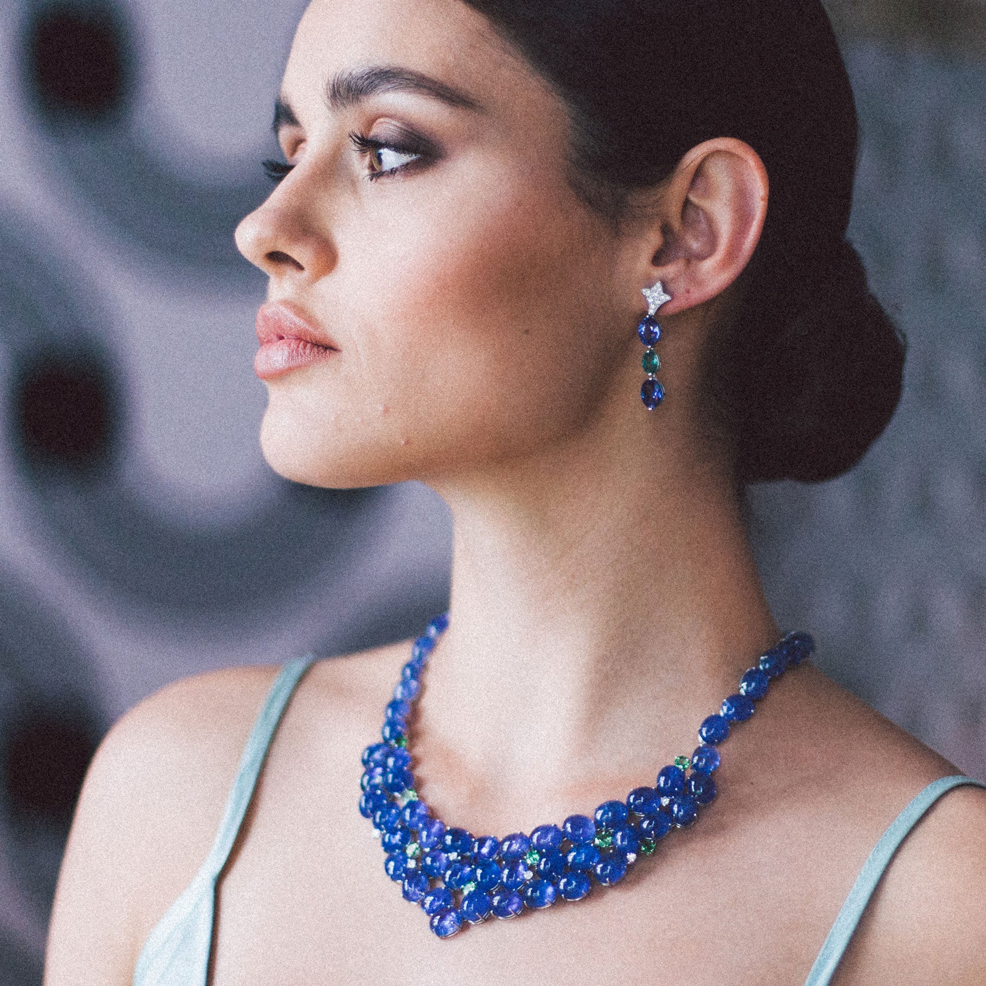 Eine Frau mit dunklem Haar blickt zur Seite. Sie trägt eine blaue Perlenkette und TANSANIT OHRGEHÄNGE von Schullin in Weißgold. Ihr Gesichtsausdruck ist ruhig, da sie ein helles Oberteil trägt. Der sanft verschwommene Hintergrund erzeugt kreisförmige Muster, die ihre Tansanit-Akzente subtil hervorheben.