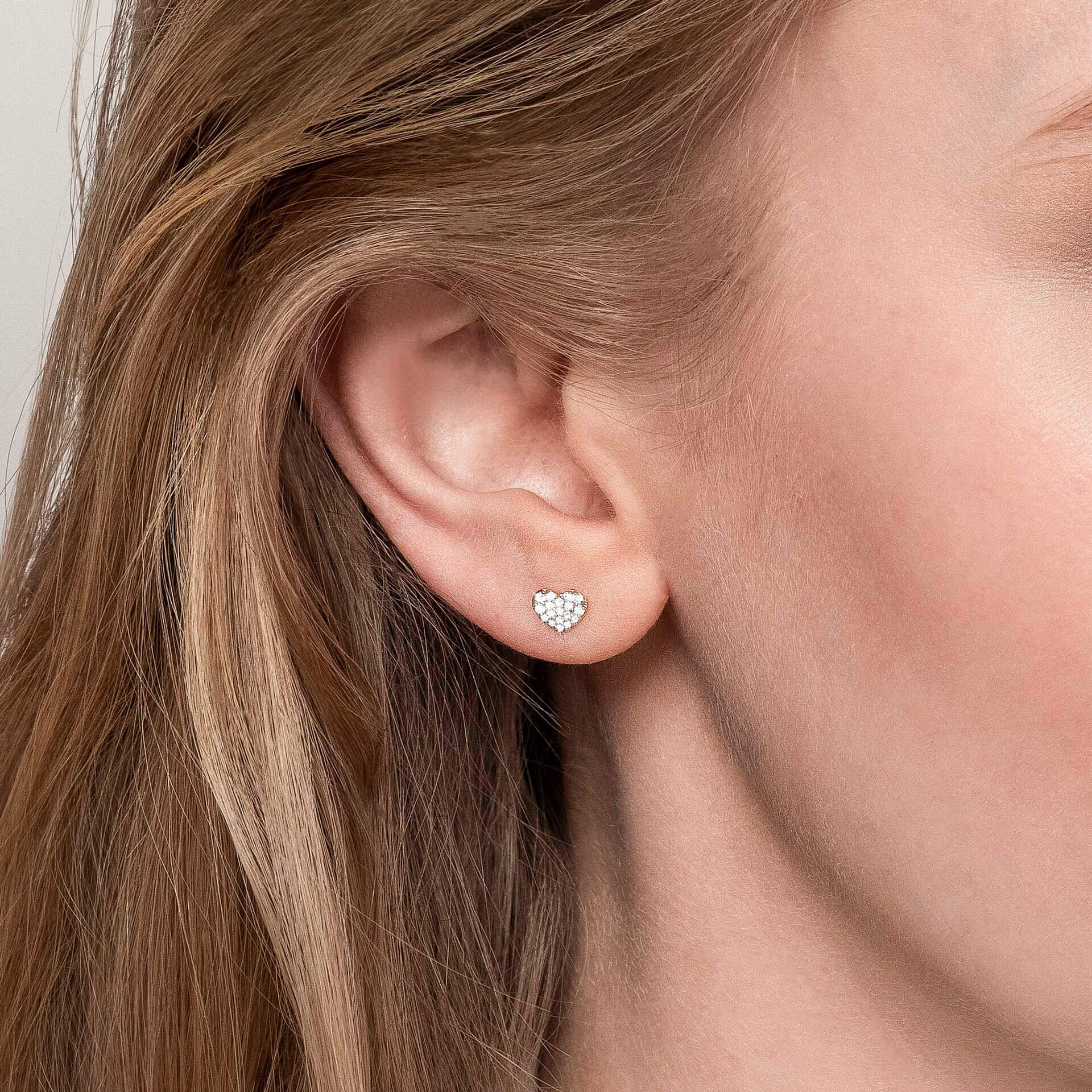 Eine Nahaufnahme zeigt einen kleinen HERZ-OHRSTECKER von Schullin, gefertigt aus 18 Karat Roségold und mit Diamanten verziert, der das Ohr einer Person ziert. Das hellbraune Haar der Person ist vor einem neutralen Hintergrund ordentlich hinter das Ohr gesteckt.