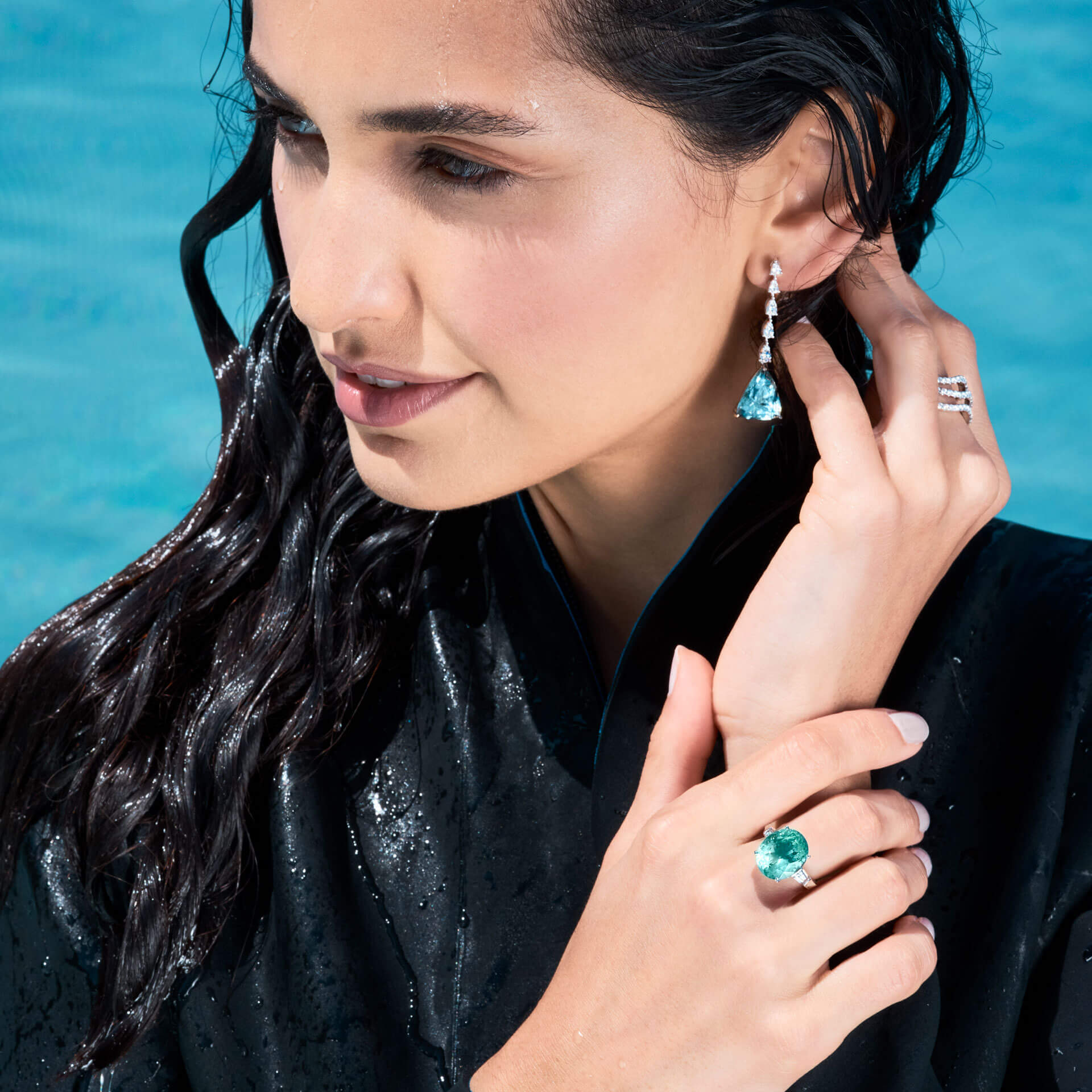 Eine Frau mit nassen Haaren, in einem dunklen Ensemble am Pool, hebt einen PARAIBA TURMALIN RING von Schullin aus 18-karätigem Weißgold sowie passende Ohrringe hervor. Mit einer Hand berührt sie sanft ihren Ohrring und mit der anderen zeigt sie prominent den glänzenden Ring. Sie steht vor einem leuchtend blauen Hintergrund.