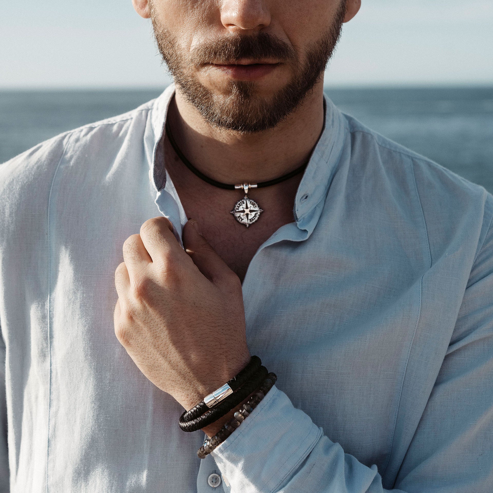 Ein bärtiger Mann steht am Meer und trägt ein hellblaues Hemd. Er berührt einen silbernen Anhänger an seiner ELEMENTS KETTE STERLING SILBER von Schullin, der die Sterling-Silber-Töne ergänzt. Mit mehreren Armbändern am Handgelenk blickt er unter einem klaren Himmel auf das Meer.