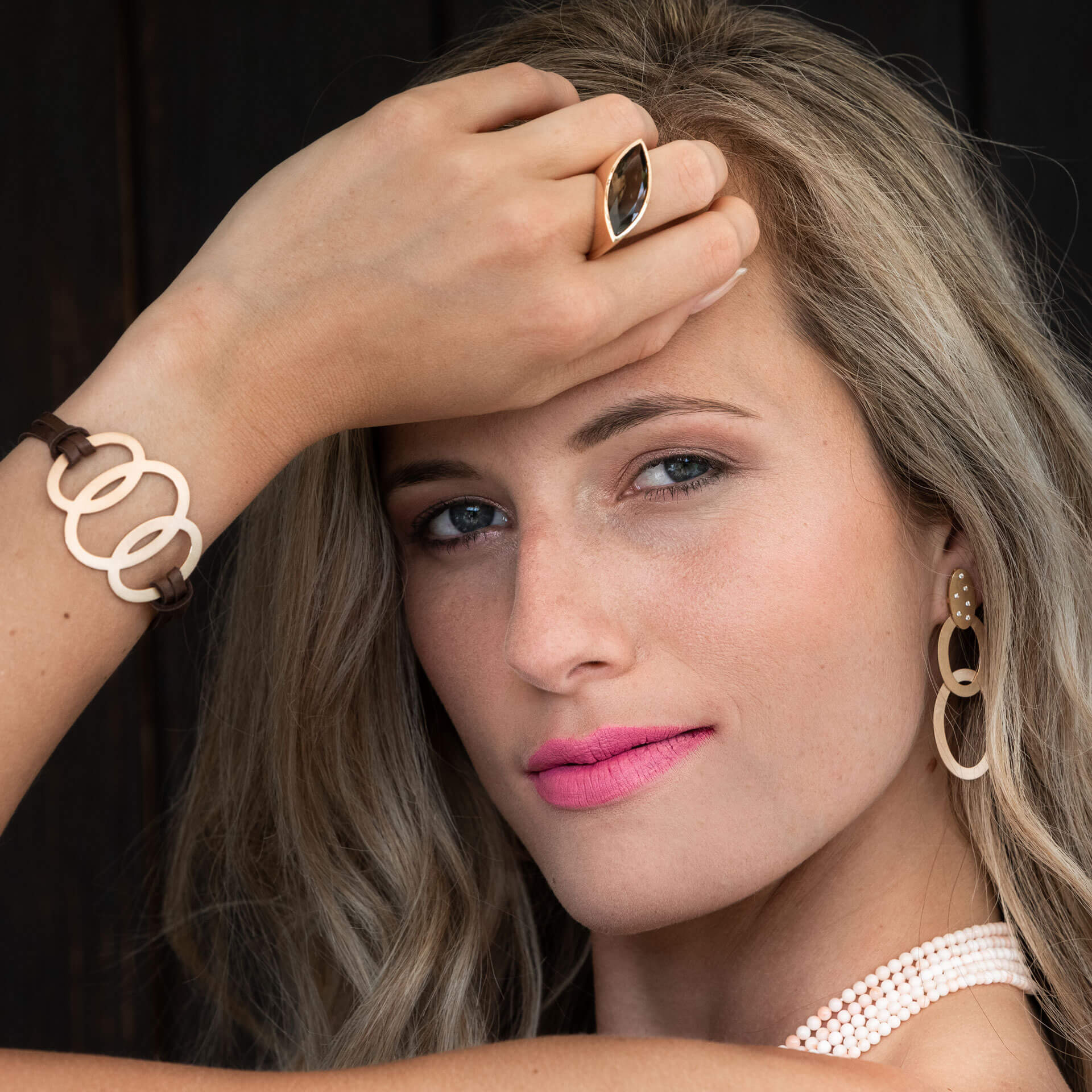 Eine Frau mit blondem Haar posiert nachdenklich vor einem dunklen Hintergrund. Ihre Hand ziert Schullins schimmerndes LEDERARMBAND ROSÉGOLD. Sie kombiniert es elegant mit runden Ohrringen und einem passenden Armband, alles hervorgehoben durch ihren rosa Lippenstift.