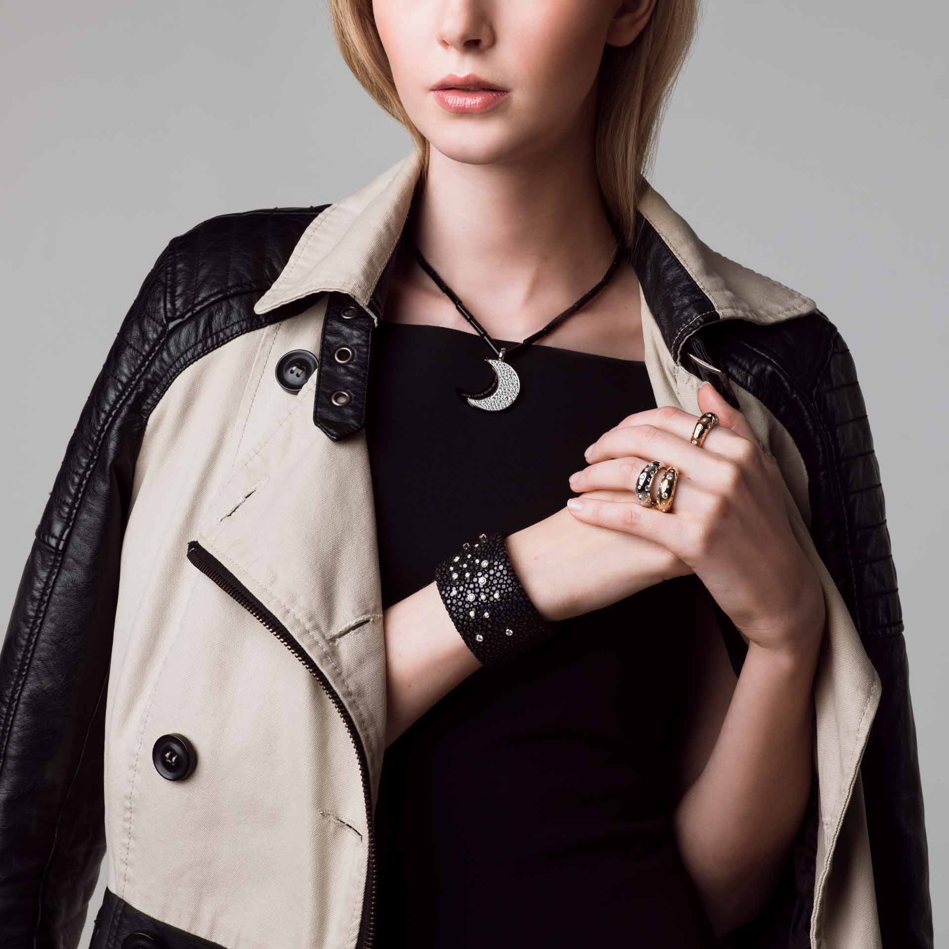 Eine Frau in einer hellen und dunklen Lederjacke und einem schwarzen Oberteil hält elegant ihr Handgelenk und präsentiert mehrere filigrane Ringe und ein Perlenarmband. Eine Halskette mit Halbmondanhänger aus Roségold verleiht ihr einen Hauch von Charme, während ihr Ensemble durch den STARDUST RING von Schullin noch aufgewertet wird. Der neutrale graue Hintergrund unterstreicht ihre stilvolle Präsenz.