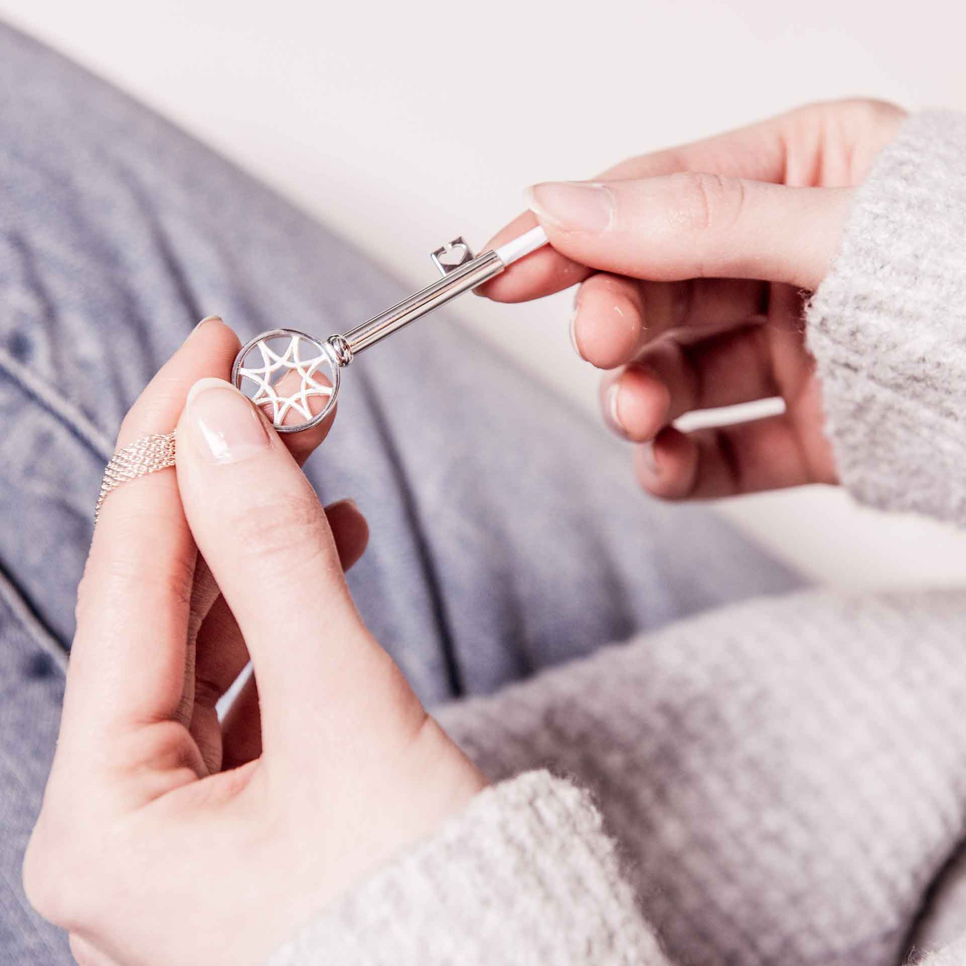 Eine Person hält den eleganten SCHLÜSSEL ZUM GLÜCK KETTE & BUCH von Schullin, der aus Sterlingsilber gefertigt ist und ein geometrisches Sternenmuster auf dem Griff aufweist. Sie trägt einen gemütlichen grauen Pullover und Jeans und konzentriert sich intensiv auf das komplizierte Design des Schlüssels.