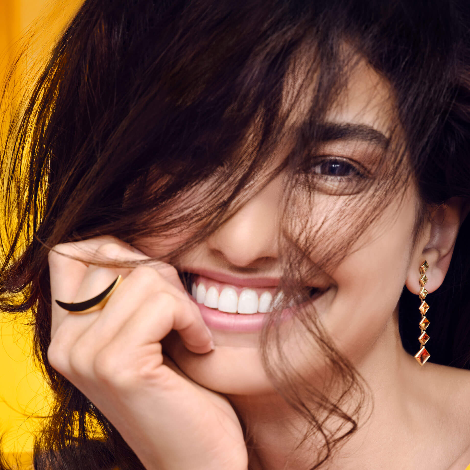 Eine Frau mit langem, dunklem Haar lächelt strahlend, ihr Gesicht ist teilweise verdeckt. Sie trägt selbstbewusst einen schwarzen GELBGOLD RING SMILE von Schullin und baumelnde rote Ohrringe. Der Hintergrund ist in einem leuchtenden Gelb gehalten und spiegelt ihre positive Lebenseinstellung wider.