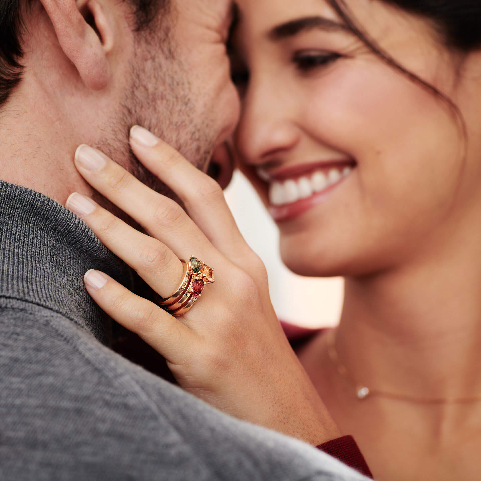 Eine Nahaufnahme eines lächelnden, sich innig umarmenden Paares hebt die Frau hervor, die elegante Ringe trägt, darunter den Schullin TURMALIN RING FANCY T mit gelbem Turmalin in 18 Karat Gelbgold an ihrer linken Hand. Ihre Hand ruht sanft auf der Schulter des Mannes. Beide wirken glücklich und sind in legere, bequeme Kleidung gekleidet. Das Bild vermittelt Wärme und Zuneigung.