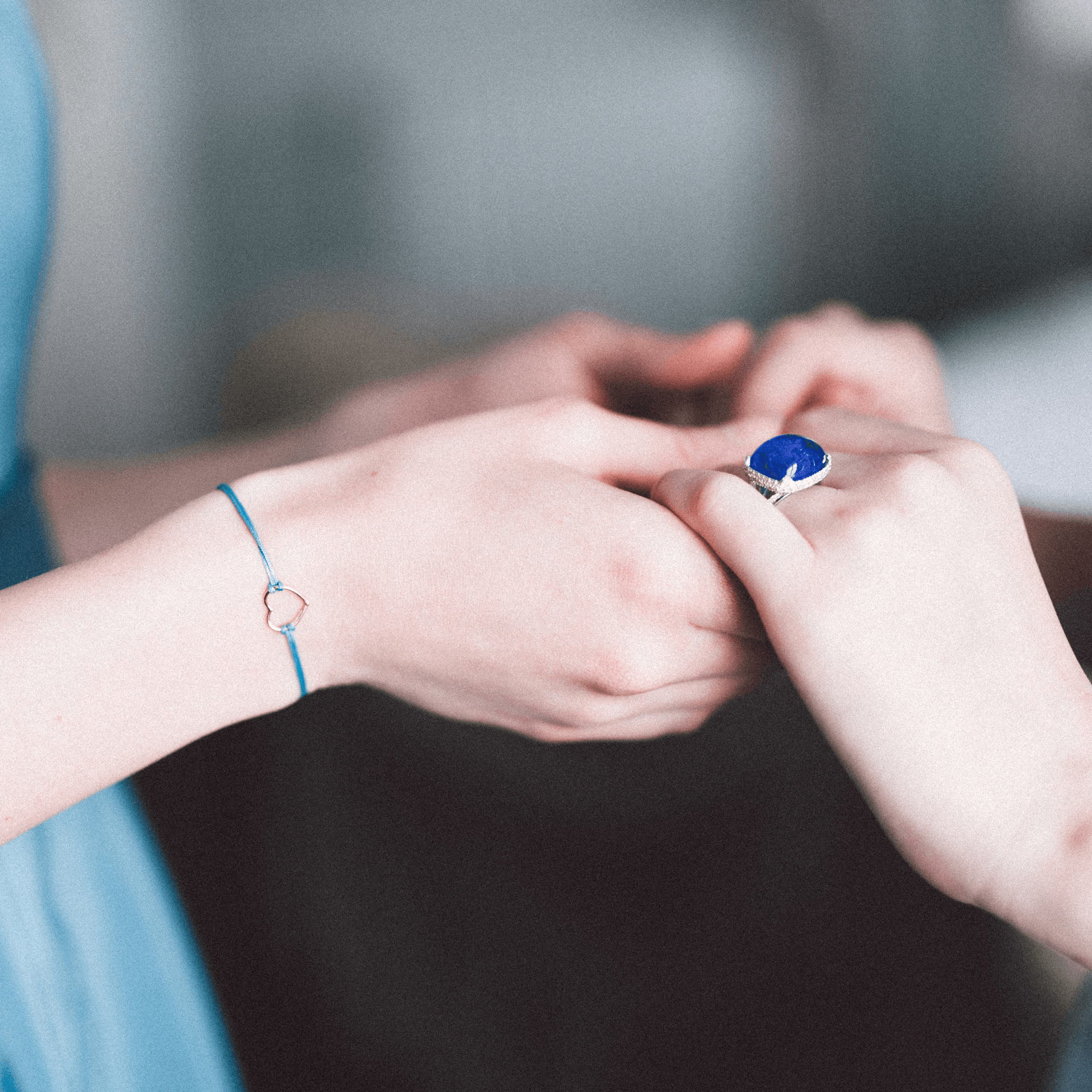Zwei Menschen mit heller Haut halten sich sanft an den Händen. Die eine trägt das HERZ ARMBAND von Schullin, ein zartes Armband mit einem kleinen Anhänger, die andere einen silbernen Ring mit einem großen blauen Stein, der vor einem sanft verschwommenen Hintergrund gefasst ist.