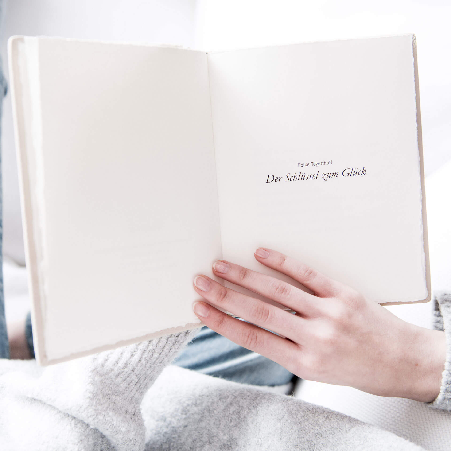 Eine Person hält den aufgeschlagenen „SCHLÜSSEL ZUM GLÜCK KETTE & BUCH“ von Schullin auf einer weißen Seite. Daneben liegt ein handgefertigter Schlüssel, der den Charme des weichen, hellen Stoffes noch verstärkt und eine ruhige, behagliche Atmosphäre verbreitet.