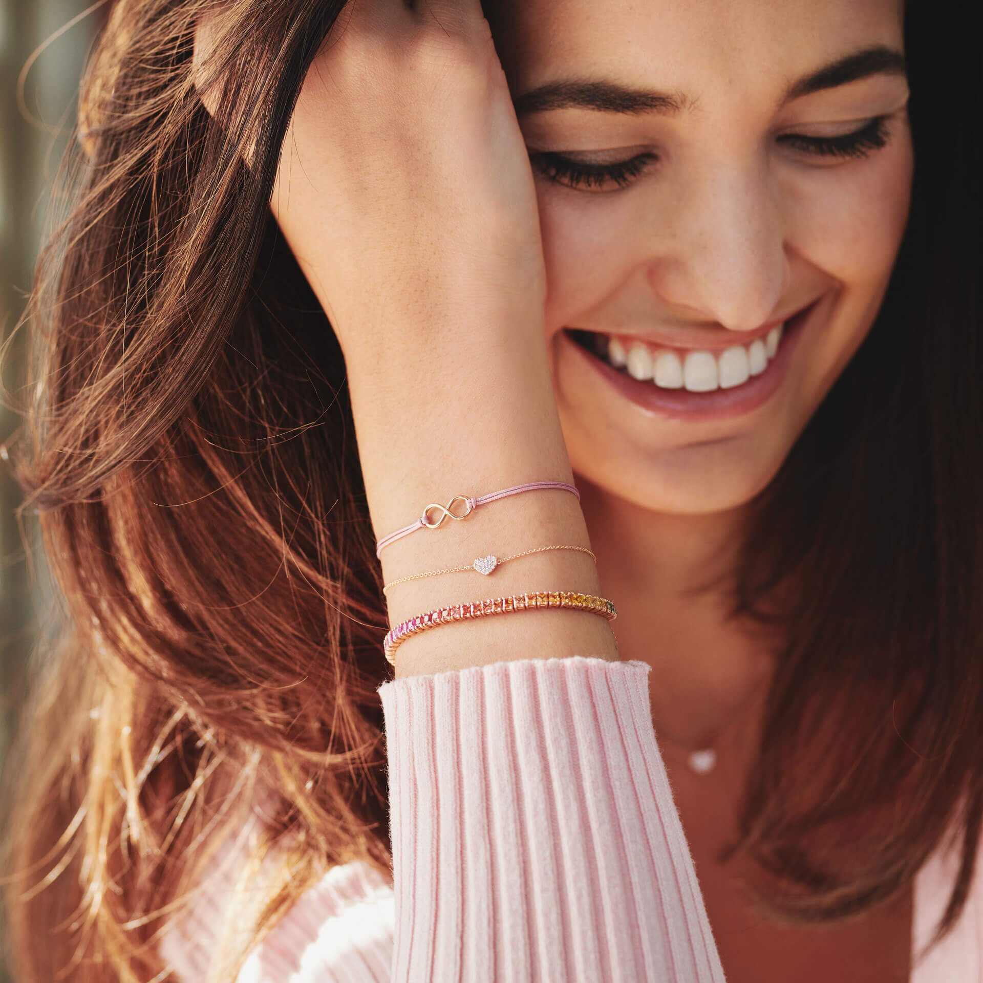 Eine Frau mit langen braunen Haaren lächelt und fasst sich an den Kopf. Sie trägt ein hellrosa Oberteil und mehrere Armbänder, darunter das zarte HERZ ARMBAND von Schullin mit einem 18 Karat Roségold-Charm, der mit Diamanten in Herzform verziert ist, sowie weitere in Gold- und Rosatönen mit Unendlichkeitssymbolen.