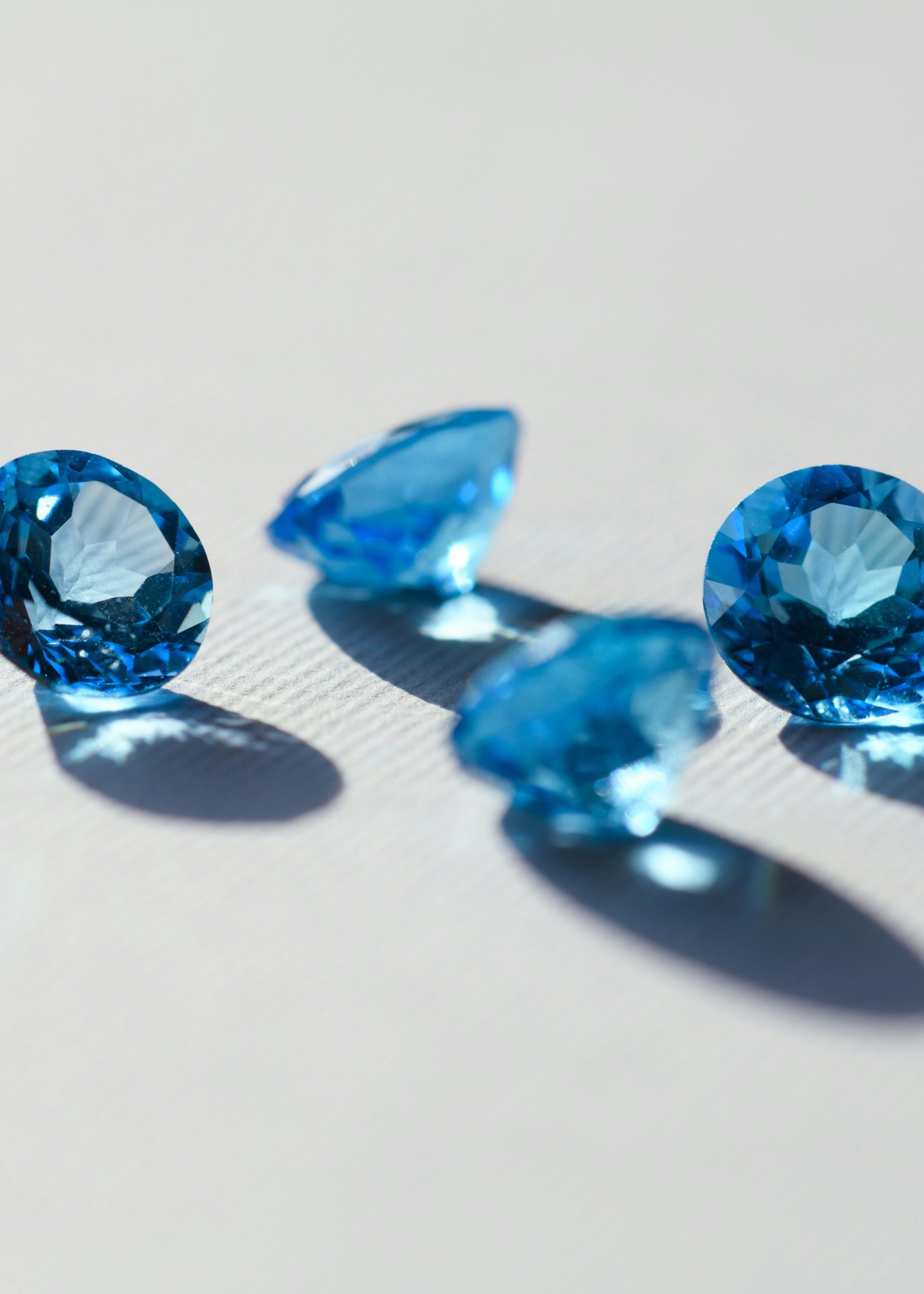 Vier blaue Edelsteine werden auf einer hellen Oberfläche präsentiert und werfen weiche Schatten. Die Edelsteine sind so geschliffen, dass sie das Licht reflektieren und ihre Facetten und leuchtenden Farben zur Geltung bringen.