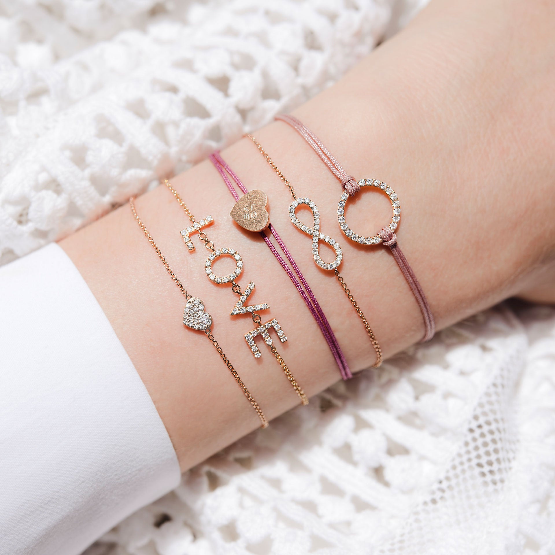 Nahaufnahme eines Handgelenks mit fünf zarten Schullin-Armbändern, darunter das HERZ ARMBAND mit Anhängern wie einem Herz, dem "LOVE"-Zeichen, dem Unendlichkeitssymbol und einem Kreis, die auf weißem, strukturiertem Stoff liegen.