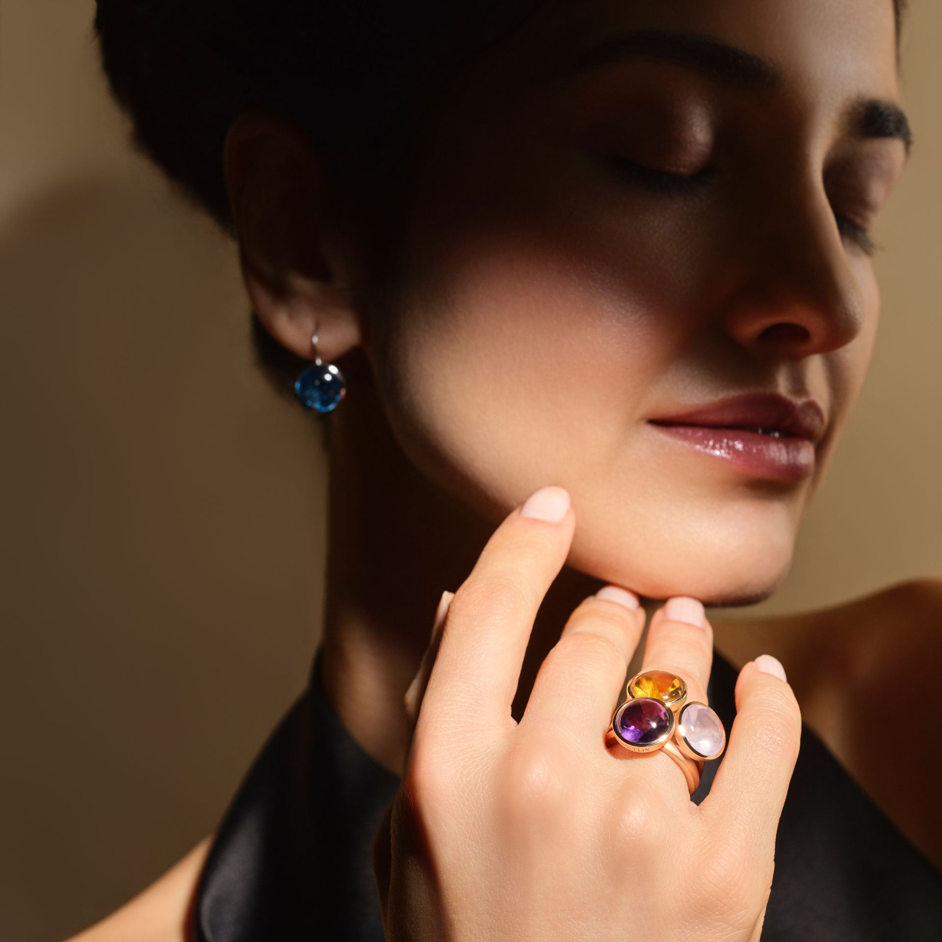 Eine Frau mit geschlossenen Augen berührt ihr Gesicht. Sie trägt ein dunkles Neckholder-Top, einen blauen Edelstein-Ohrring und den Schullin ROSENQUARZ RING BUFFTOP-Gold, besetzt mit lila, gelben und klaren Steinen. Sanftes Licht unterstreicht ihren heiteren Ausdruck.