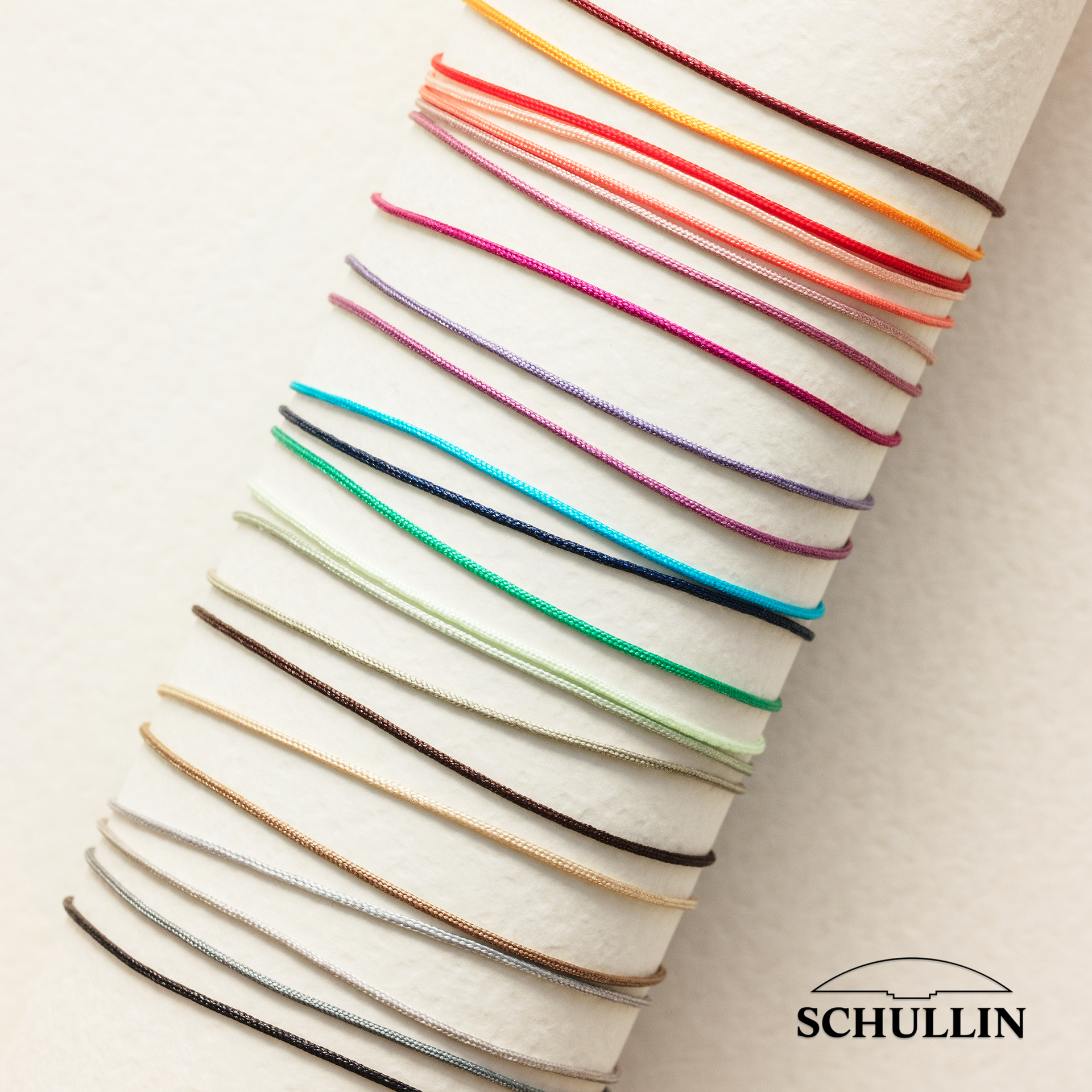 Auf einer strukturierten cremefarbenen Oberfläche sind zahlreiche Bänder in leuchtenden Farbtönen wie Rosa, Rot, Orange, Lila, Grün, Blau, Braun und Grau ordentlich angeordnet. In der Ecke ist ein Schullin-Logo.