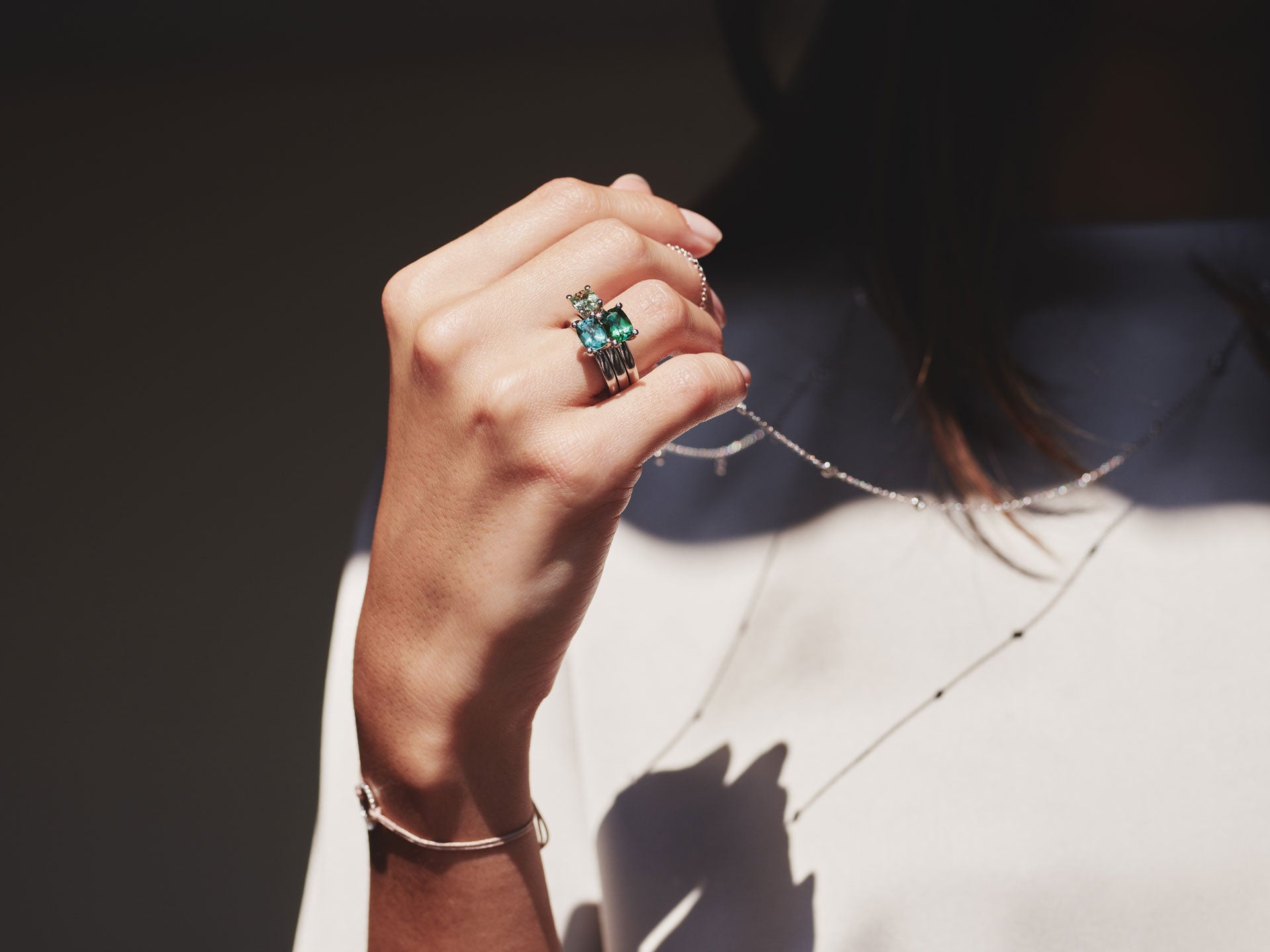 In der Hand einer Frau ist der Schullin TURMALIN RING FANCY T neben mehreren gestapelten Ringen und einem Armband zu sehen, während eine zarte Halskette und ein Teil ihres weißen Oberteils in sanfter Beleuchtung sichtbar sind.