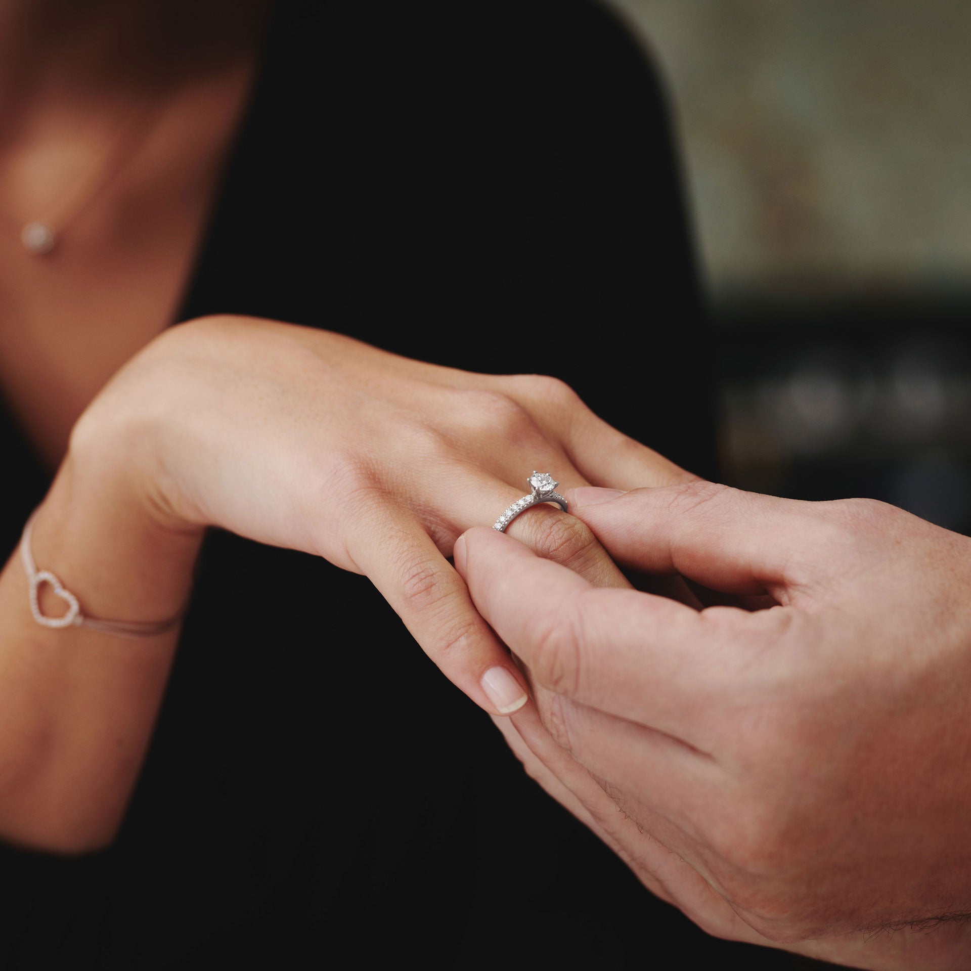 Eine Nahaufnahme zeigt eine Person, die den Verlobungsring Schullin RING GRACE 0.75 CARAT an den manikürten Finger einer anderen Person steckt, die ein herzförmiges Armband trägt, mit einem leicht verschwommenen Hintergrund.