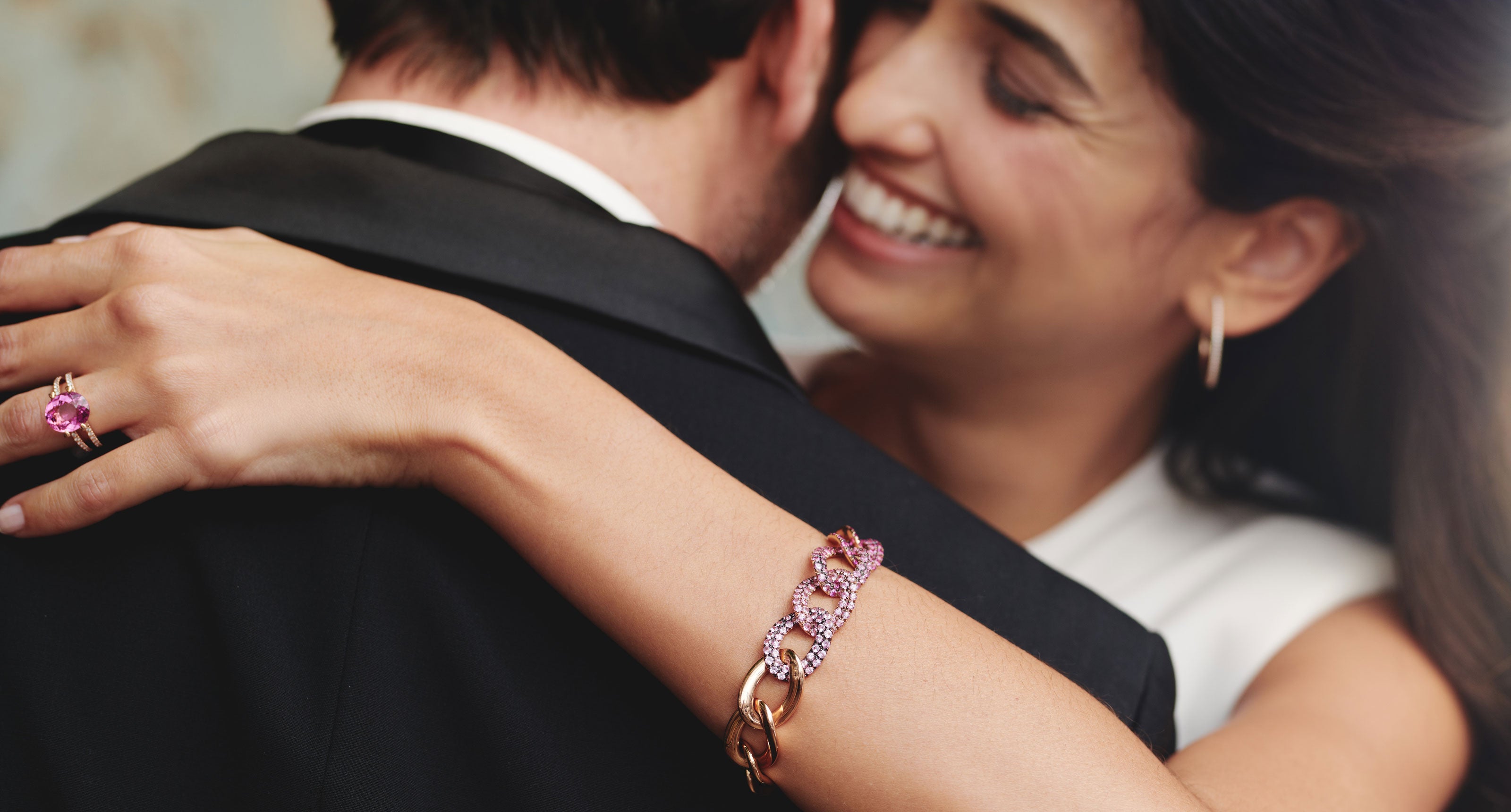 Eine lächelnde Frau umarmt einen Mann im Anzug. Sie trägt ein goldenes und rosafarbenes Armband, Ohrringe und einen großen rosafarbenen Ring und hat ihren Arm um seinen Hals gelegt. Ihre Gesichter sind nah beieinander und zeigen Zuneigung und Glück.