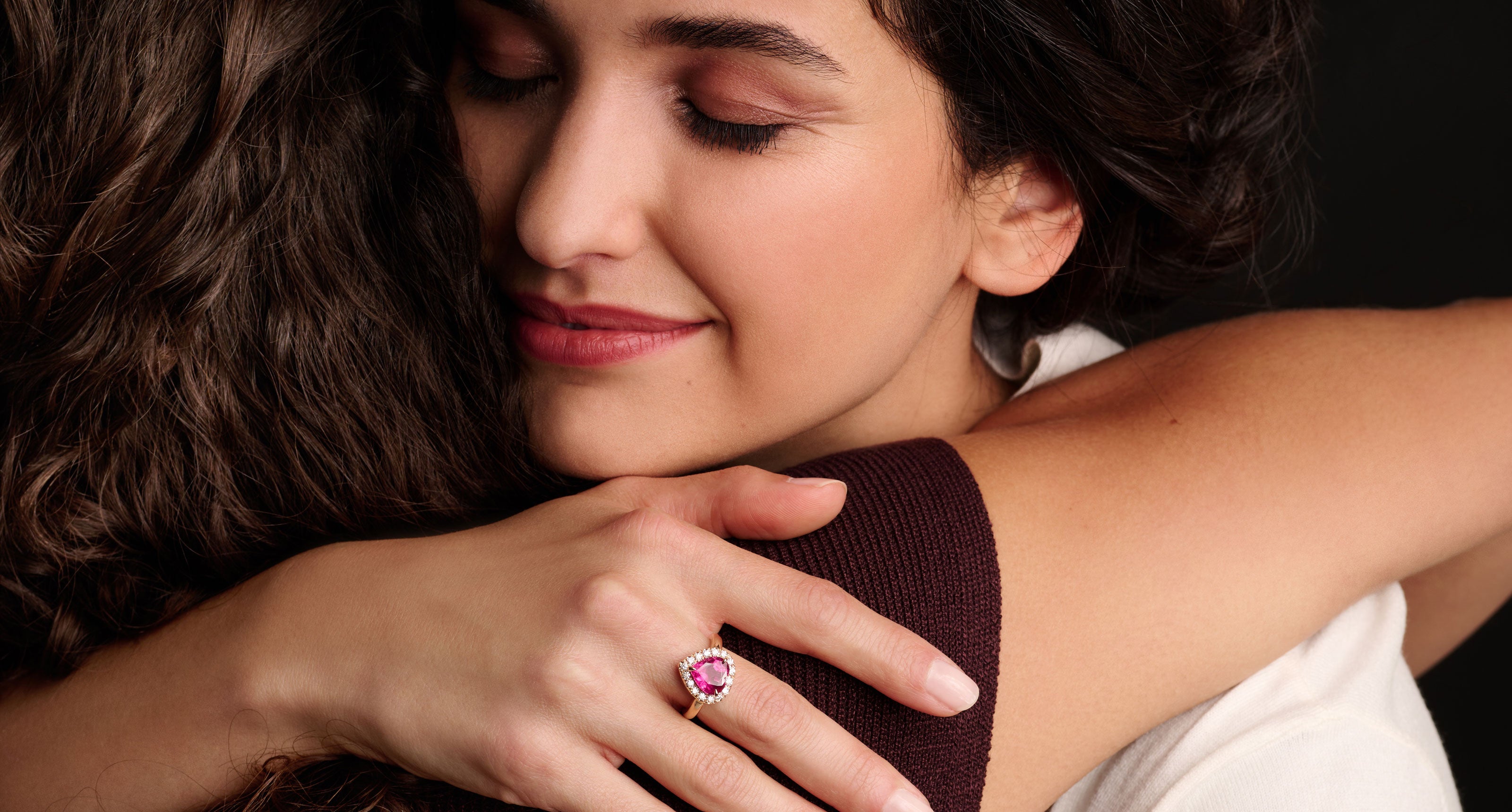 Eine Frau mit geschlossenen Augen lächelt sanft, während sie eine andere Person umarmt. Sie trägt einen Ring mit einem rosafarbenen herzförmigen Stein an ihrem Finger. Der Hintergrund ist dunkel und unterstreicht die Wärme und Nähe der Umarmung.