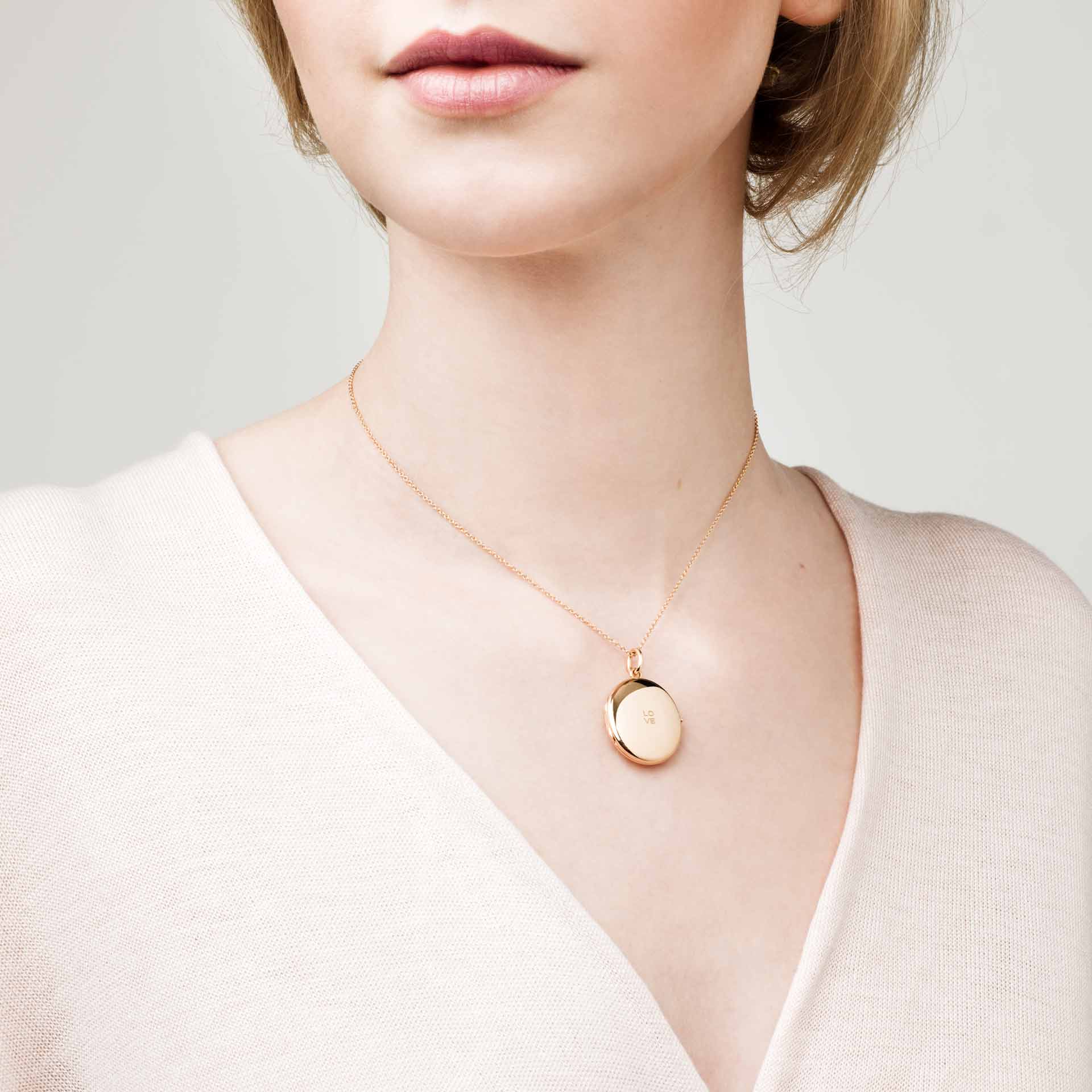 Eine Frau trägt das goldene Medaillon Schullin MEDAILLON LOVE an einer zarten Kette, kombiniert mit einem hellrosa Oberteil mit V-Ausschnitt. Das Bild hebt ihren Hals und ihre obere Brust hervor, wobei das runde Medaillon auf ihrer Haut zu sehen ist.