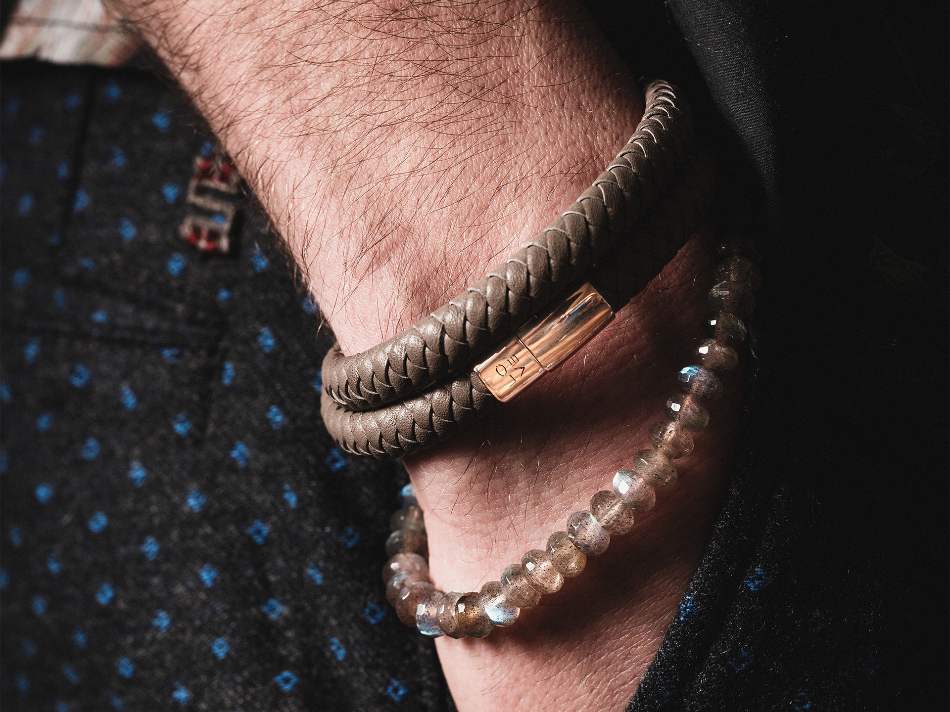 Nahaufnahme eines mit zwei Armbändern geschmückten Handgelenks: Schullins POWER ARMBAND MIT GRAVUR, ein geflochtenes Lederband mit Metallschließe, und eine Kette aus durchsichtigen Perlen. Die Trägerin trägt eine dunkel gemusterte Hose.